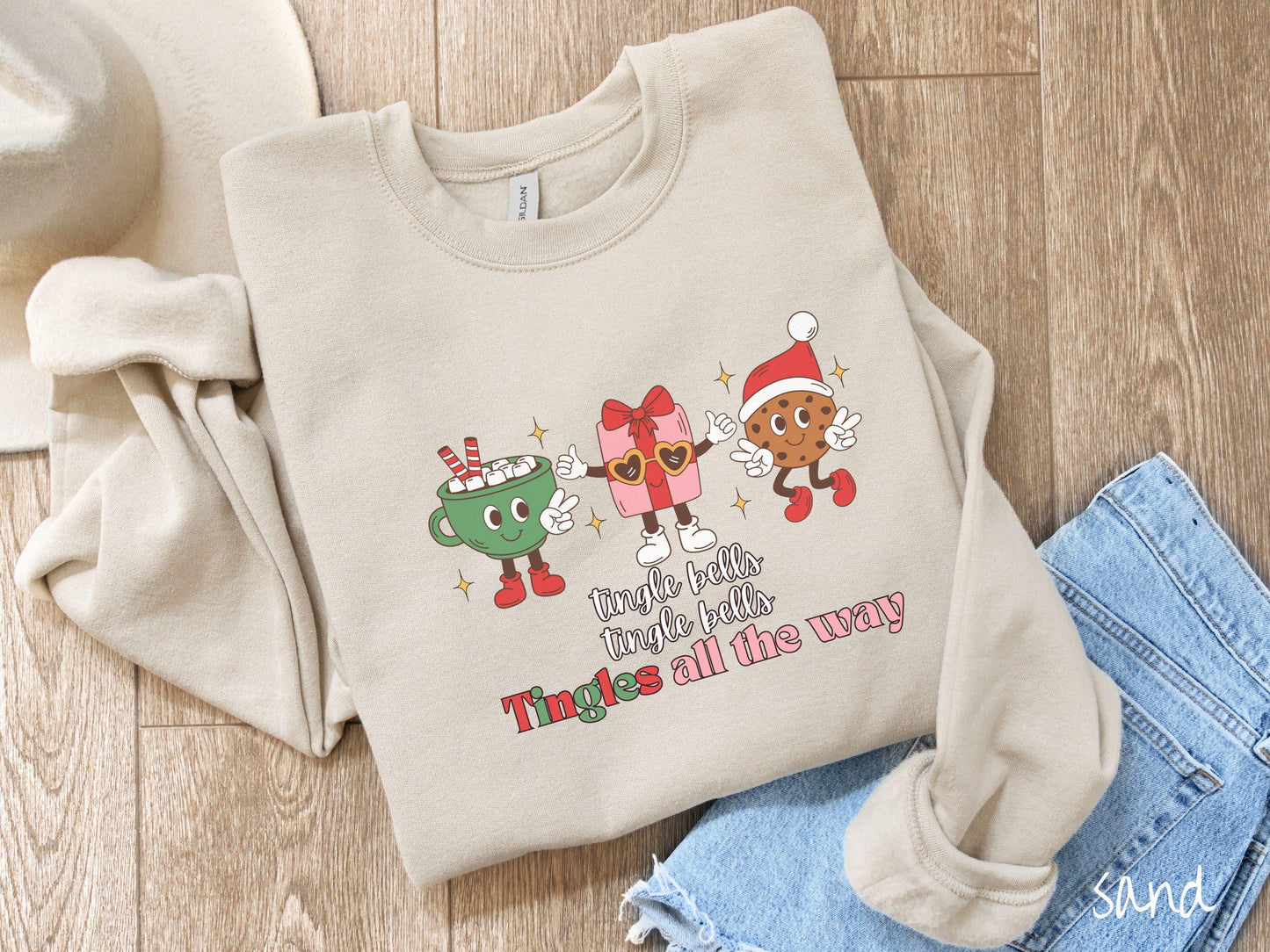 A cute, vintage sand colored Gildan sweatshirt with text tingle bells, tingle bells, Tingles all the way. Above are a green cup of hot cocoa, a red and pink present wearing heart-shaped glasses, and a chocolate chip cookie wearing a Santa hat.