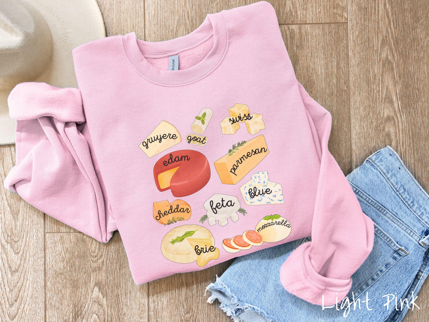 A vintage light pink colored Gildan sweatshirt is perfect for cheese lovers with a grid pattern of various cheeses like feta, parmesan, gruyere, goat, swiss, edam, cheddar, blue, brie, and mozzarella listed.