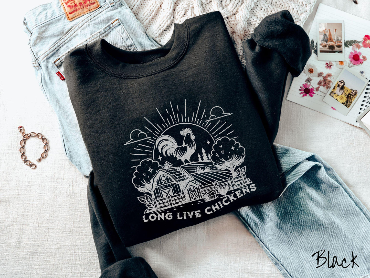 A vintage black colored Gildan sweatshirt featuring a design of a farm with a fence, trees, and plants scattered along the property, a huge chicken in the background at the top of a pasture in front of the sun, and text Long Live Chickens.