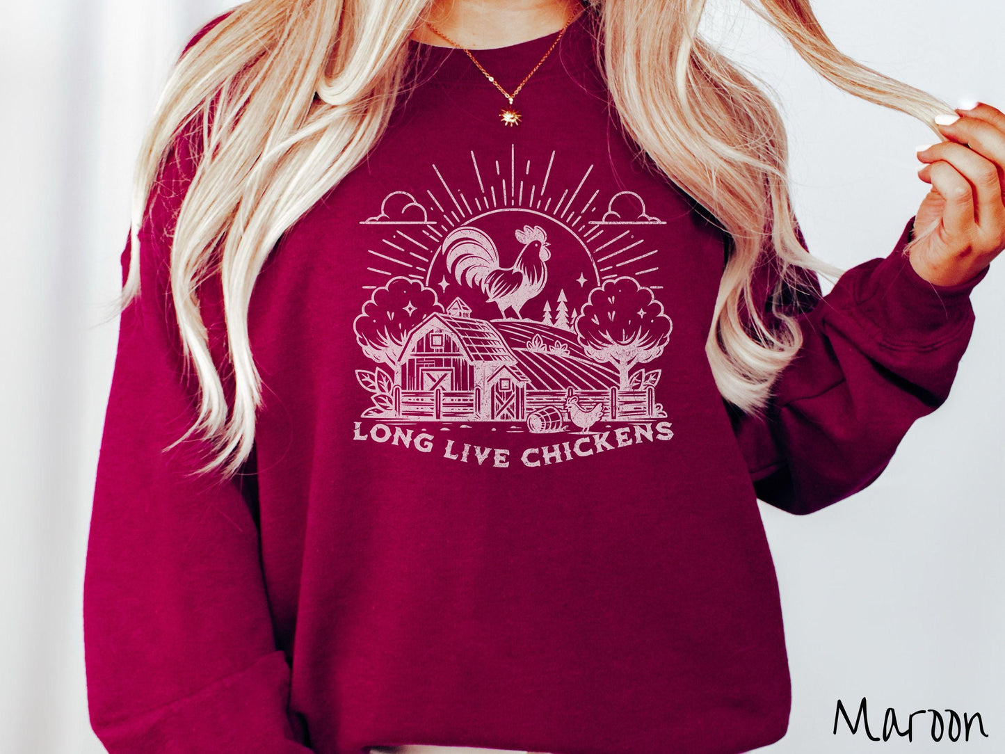 A vintage maroon colored Gildan sweatshirt featuring a design of a farm with a fence, trees, and plants scattered along the property, a huge chicken in the background at the top of a pasture in front of the sun, and text Long Live Chickens.