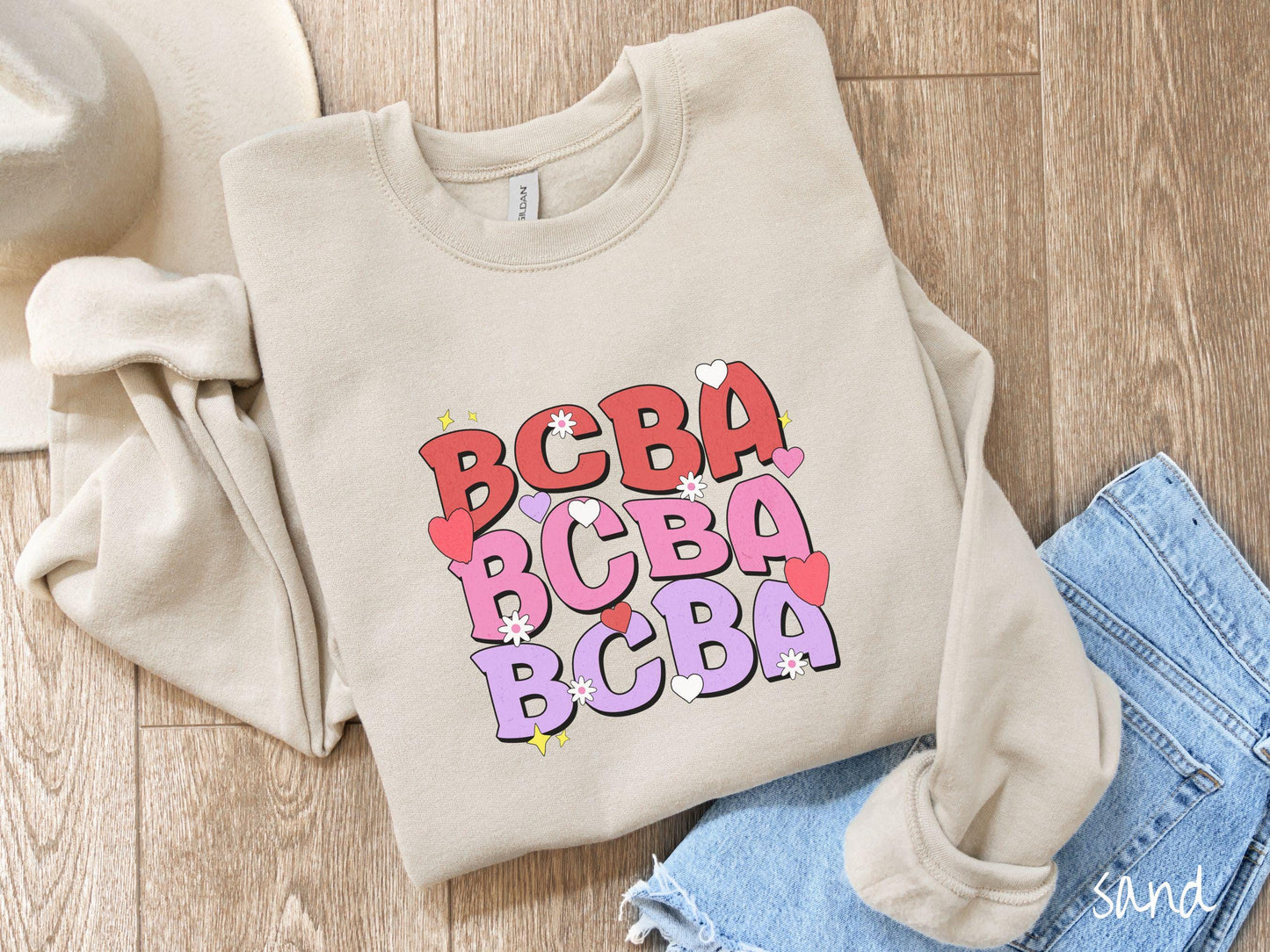 A vibrant Gildan Sweatshirt with the text BCBA written three times vertically, accompanied by tiny hearts, stars, and flowers. Perfect for Board Certified Behavior Analysts, this tee is a great Valentine's Day gift to show appreciation and love.