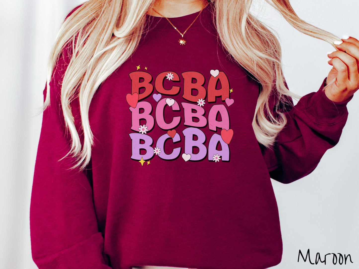 A vibrant Gildan Sweatshirt with the text BCBA written three times vertically, accompanied by tiny hearts, stars, and flowers. Perfect for Board Certified Behavior Analysts, this tee is a great Valentine's Day gift to show appreciation and love.