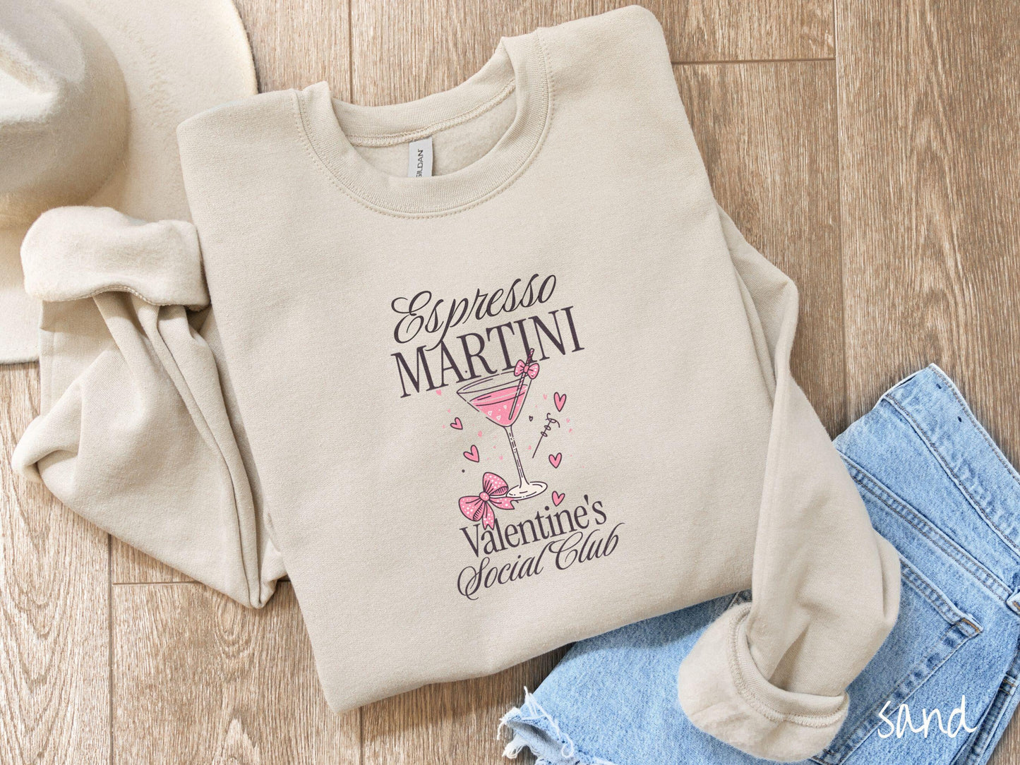 A stylish Gildan sweatshirt featuring the phrase "Espresso Martini" above a martini glass filled with pink martini and a pink bow-topped toothpick. The words "Valentine's Social Club" appear below with pink bows and hearts throughout the design.