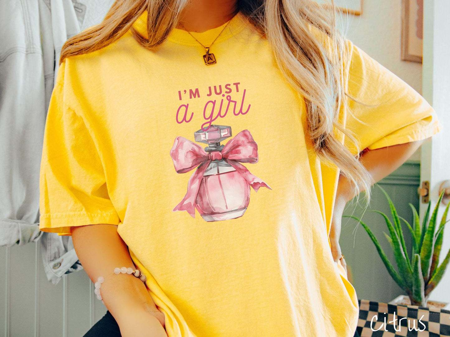 A stylish Comfort Colors T-shirt featuring the phrase "I'm Just a Girl" in pink font above a bottle of pink perfume tied with a pink bow. This tee exudes a feminine and trendy vibe, perfect for those who appreciate unique fashion statements.