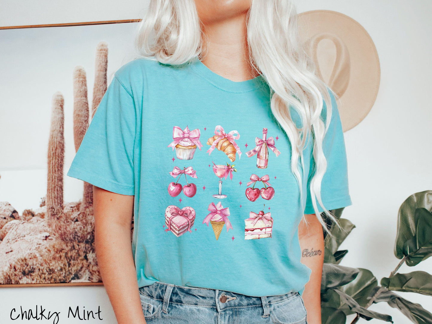 A stylish coquette Comfort Colors T-shirt featuring a grid of assorted treats with pink ribbons like a cupcake, a croissant, a champaigne bottle, cherries, a pink martini, a pink cake, and a pink ince cream cone, along with twinkling pink stars.