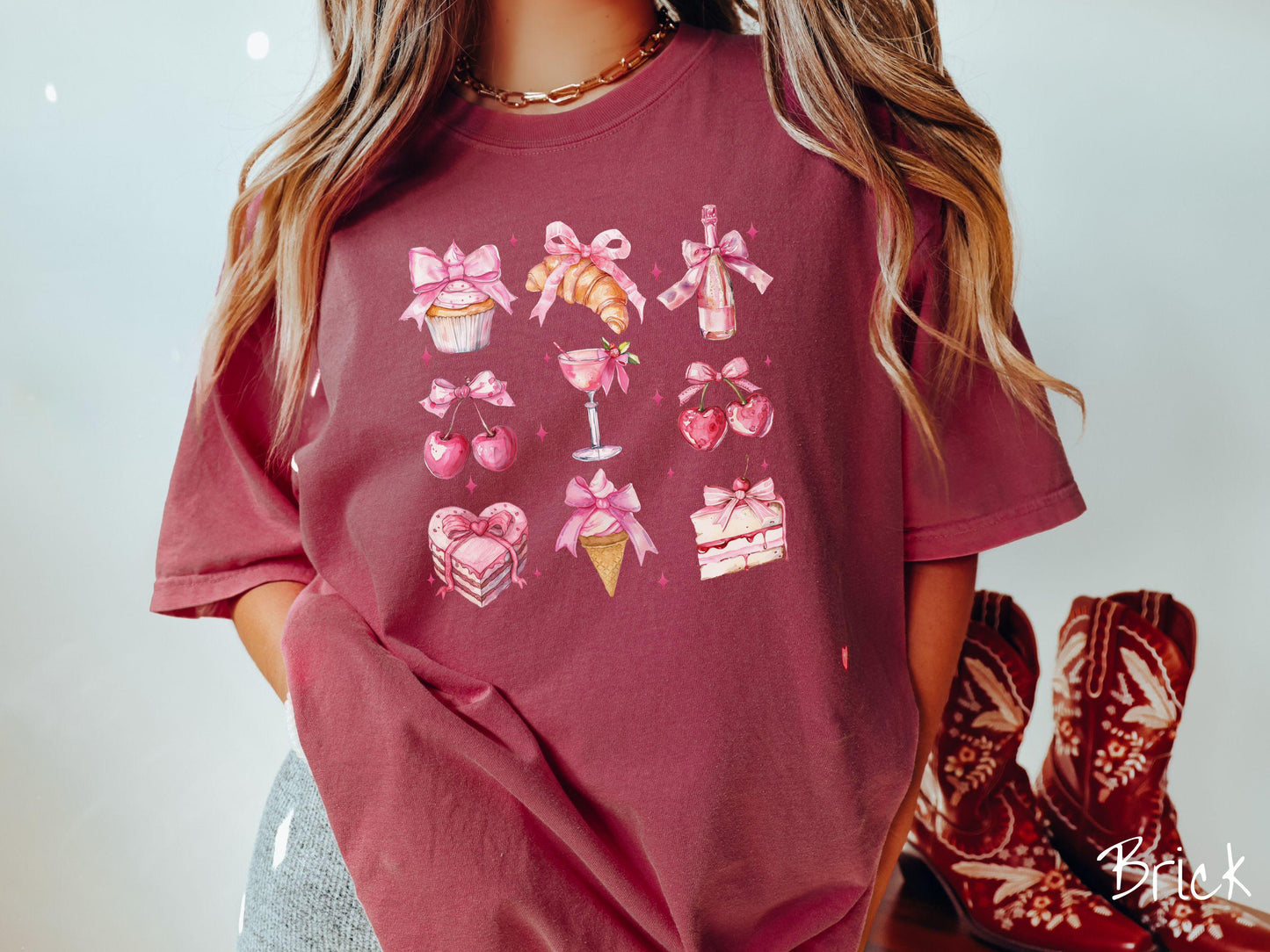 A stylish coquette Comfort Colors T-shirt featuring a grid of assorted treats with pink ribbons like a cupcake, a croissant, a champaigne bottle, cherries, a pink martini, a pink cake, and a pink ince cream cone, along with twinkling pink stars.