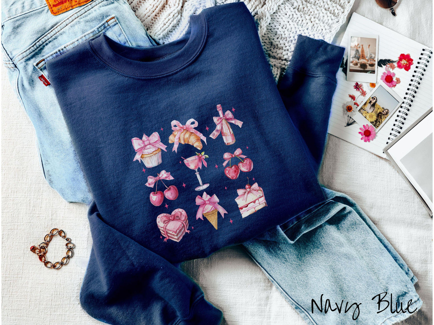 A stylish coquette Gildan sweatshirt featuring a grid of assorted treats with pink ribbons like a cupcake, a croissant, a champaigne bottle, cherries, a pink martini, a pink cake, and a pink ince cream cone, along with twinkling pink stars.