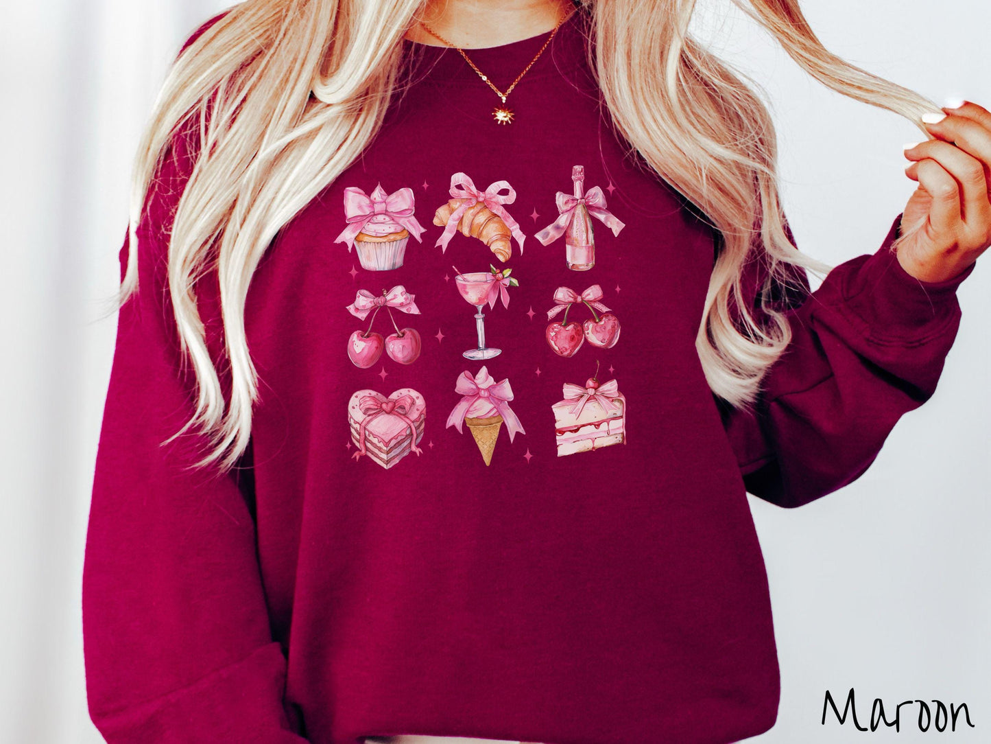 A stylish coquette Gildan sweatshirt featuring a grid of assorted treats with pink ribbons like a cupcake, a croissant, a champaigne bottle, cherries, a pink martini, a pink cake, and a pink ince cream cone, along with twinkling pink stars.