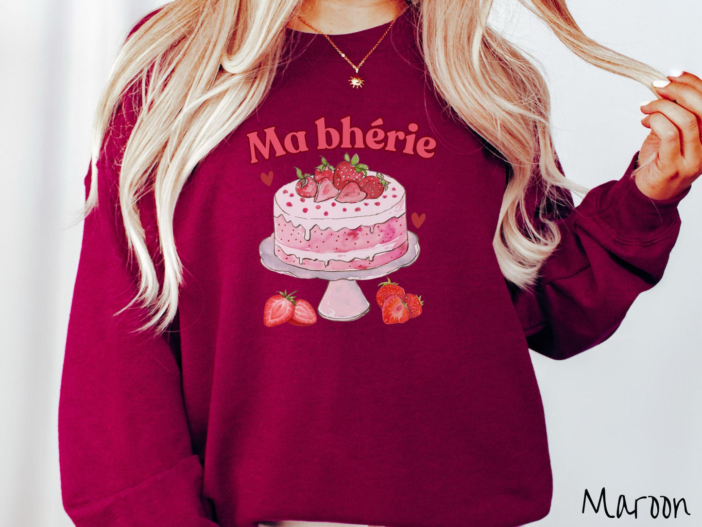 A Gildan sweatshirt featuring the phrase "Ma Bherie" is printed in bold red font above a charming illustration of a red-and-white strawberry cake displayed on a plate, topped with fresh strawberries and surrounded by even more berries.