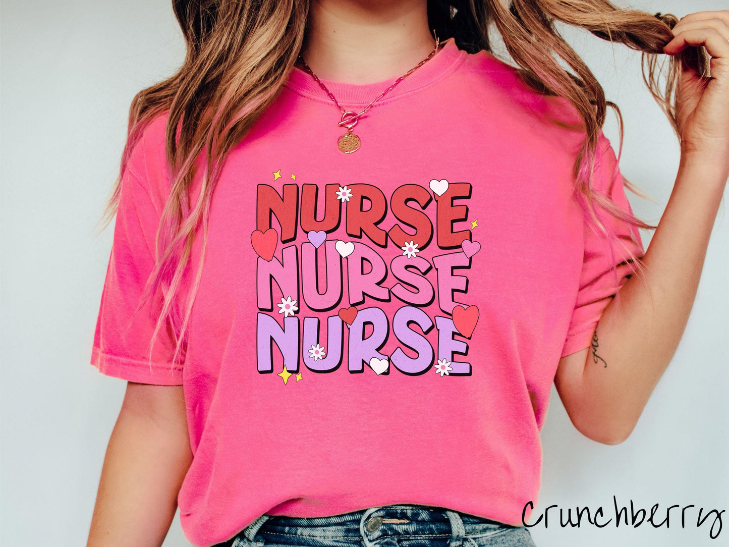 A stylish Comfort Colors T-shirt featuring the word "NURSE" written three times vertically in red, pink, and purple font. There are red, white, and purple hearts mixed into the design as well as yellow twinkling stars.