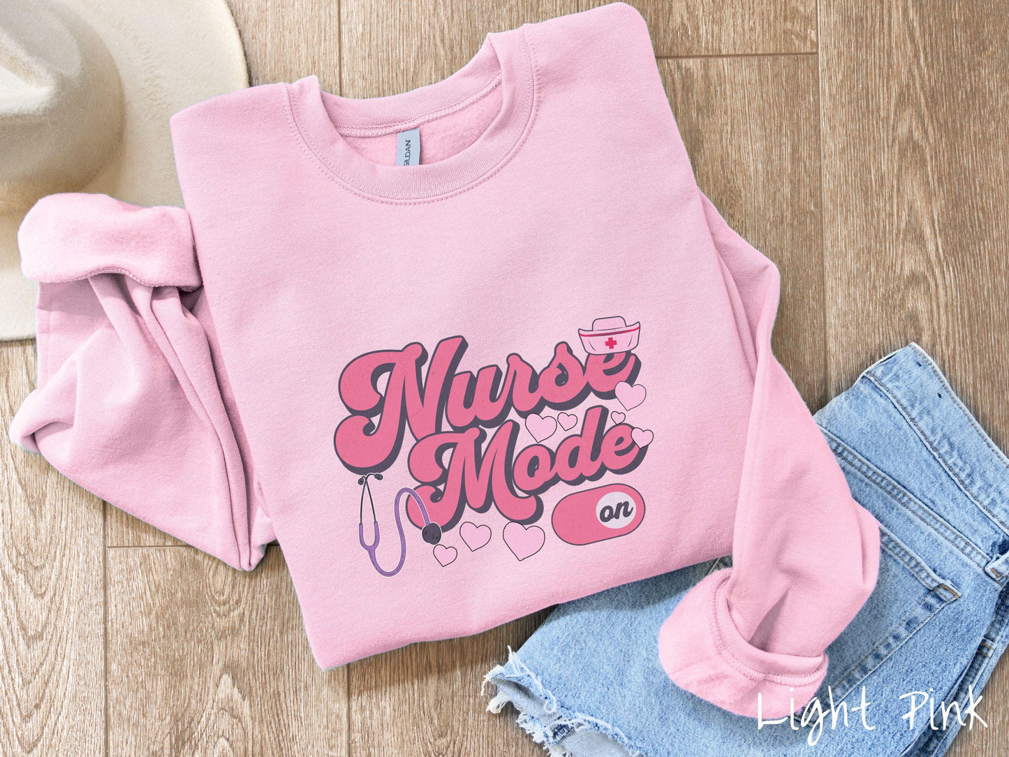 A stylish Gildan sweatshirt design features the text "Nurse Mode" in bold pink font with a pink nurse's cap placed over the letter E, and a stethoscope positioned beneath the text, alongside an "on" button widget set to the on position.