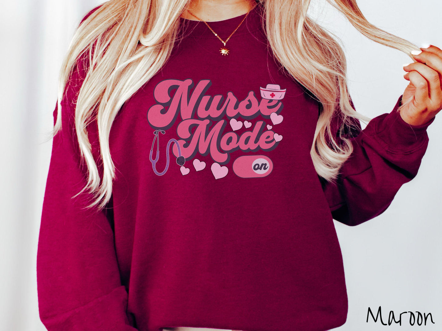 A stylish Gildan sweatshirt design features the text "Nurse Mode" in bold pink font with a pink nurse's cap placed over the letter E, and a stethoscope positioned beneath the text, alongside an "on" button widget set to the on position.