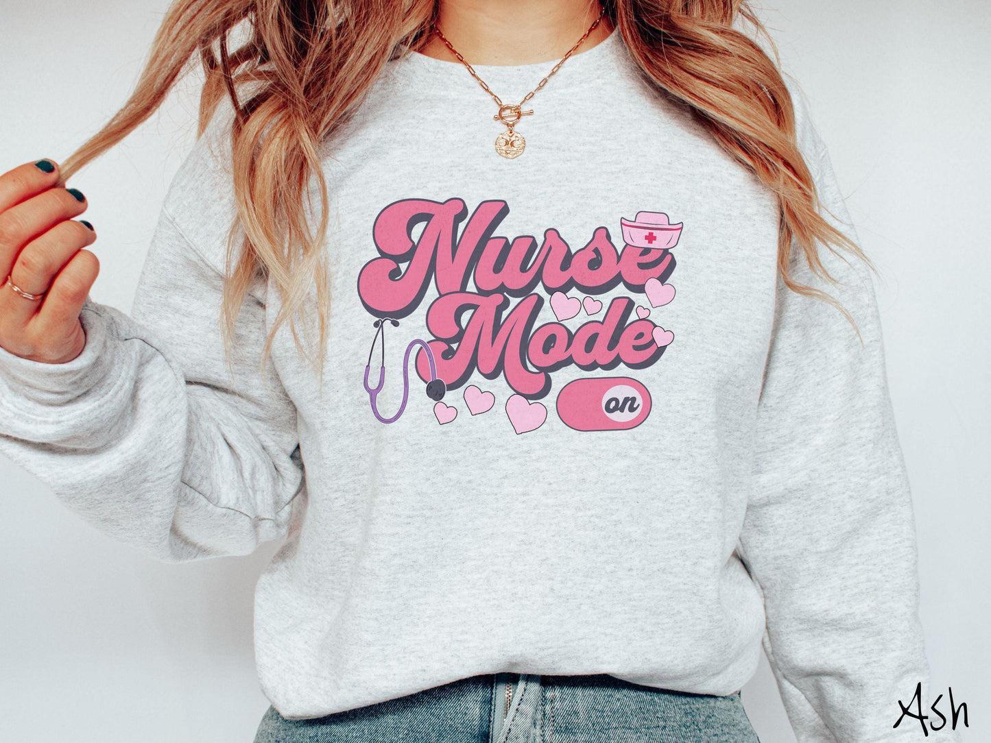 A stylish Gildan sweatshirt design features the text "Nurse Mode" in bold pink font with a pink nurse's cap placed over the letter E, and a stethoscope positioned beneath the text, alongside an "on" button widget set to the on position.