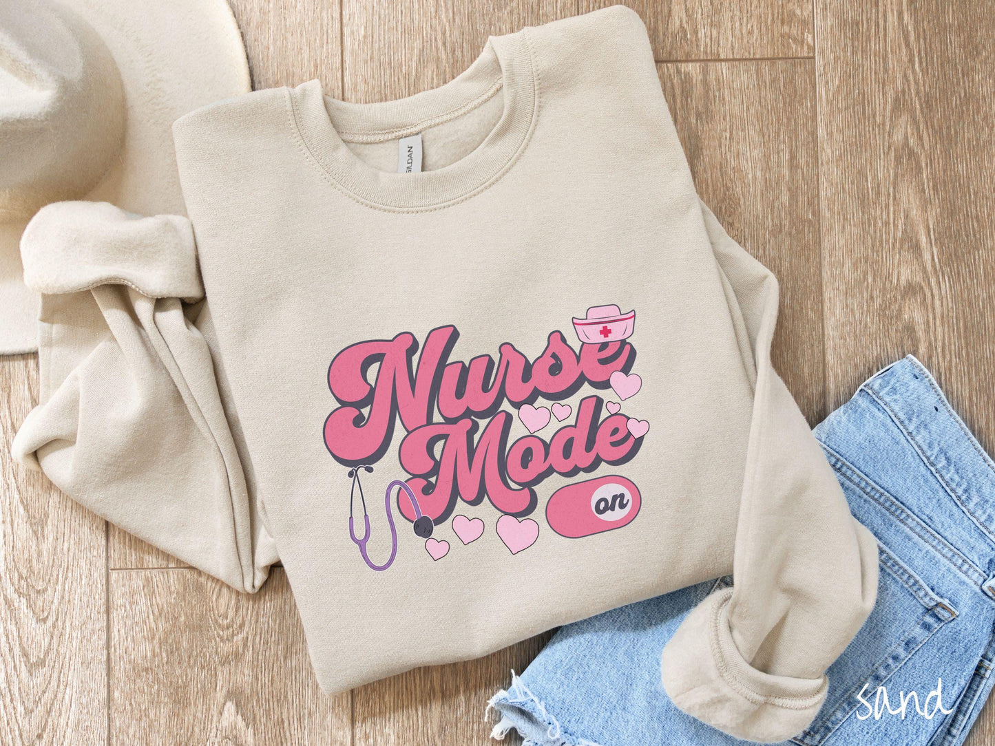 A stylish Gildan sweatshirt design features the text "Nurse Mode" in bold pink font with a pink nurse's cap placed over the letter E, and a stethoscope positioned beneath the text, alongside an "on" button widget set to the on position.