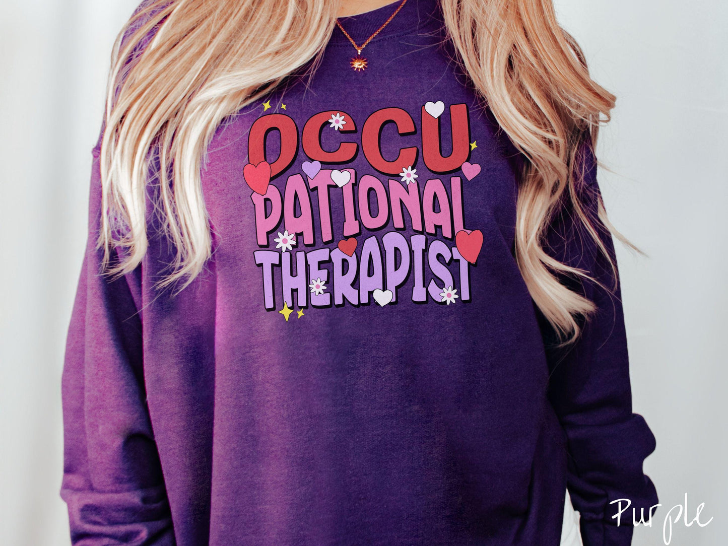 A stylish Gildan sweatshirt featuring the words "Occupational Therapist" arranged vertically in bold red, pink, and purple font, this design is enhanced with colorful hearts, flowers, and stars for a playful, uplifting touch.