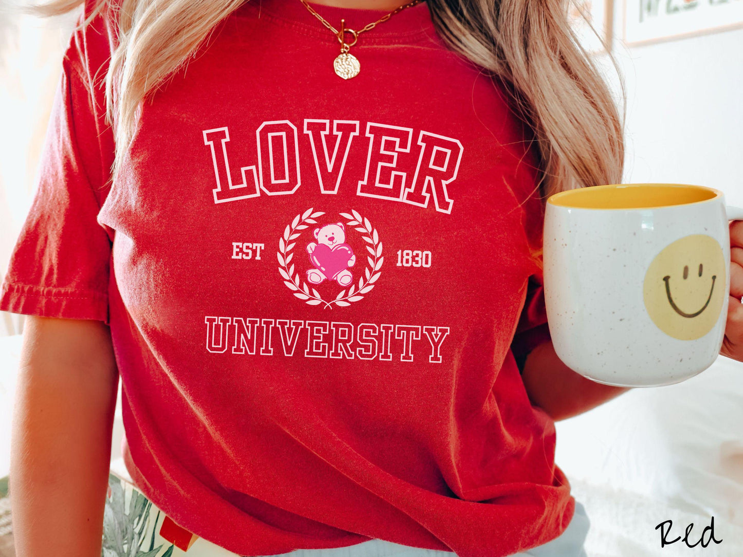 A vintage Comfort Colors t-shirt featuring a university logo-inspired design showcasing the text “Lover University” in collegiate-style font. Two pink laurel leaves frame a pink teddy bear hugging a heart with the text “EST. 1830” in pink font.