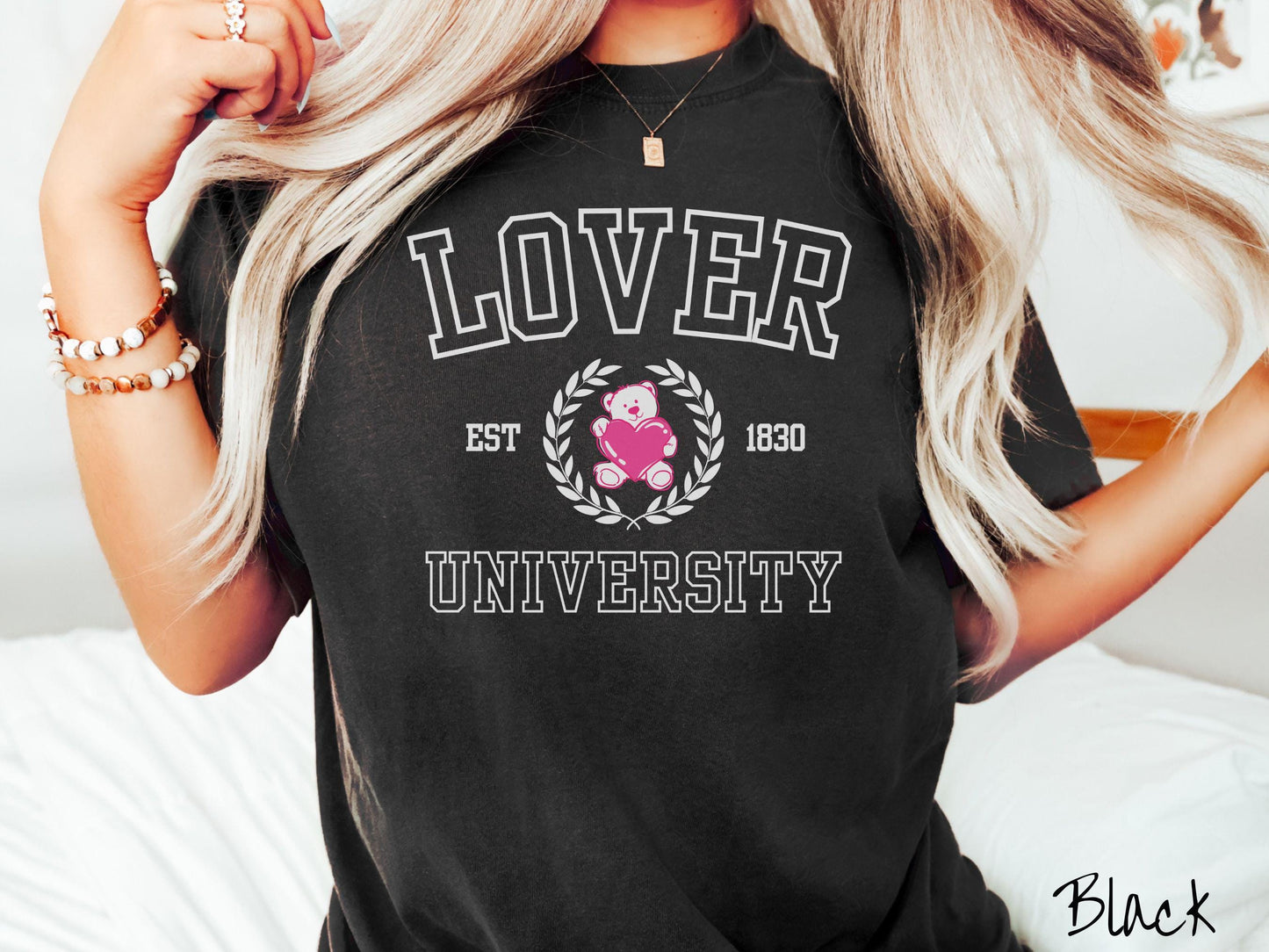 A vintage Comfort Colors t-shirt featuring a university logo-inspired design showcasing the text “Lover University” in collegiate-style font. Two pink laurel leaves frame a pink teddy bear hugging a heart with the text “EST. 1830” in pink font.
