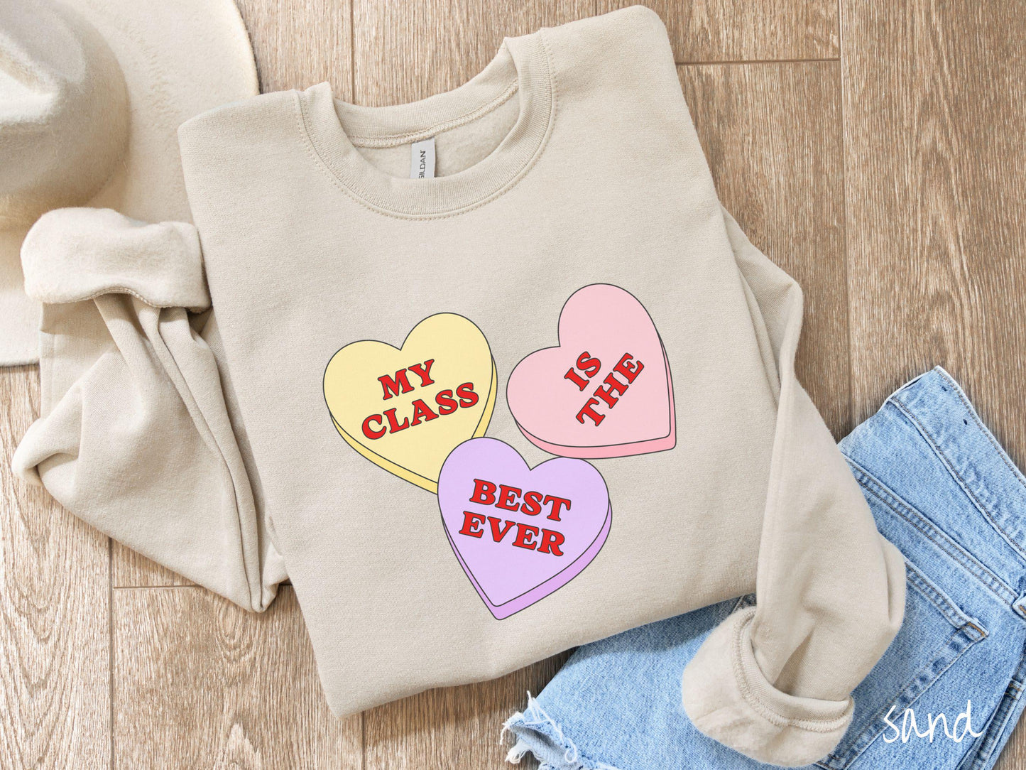 A charming vintage-style Valentine’s Day Gildan sweatshirt featuring three vibrant conversation hearts in yellow, red, and purple, this design displays the text “My class is the best ever” spread across the hearts.