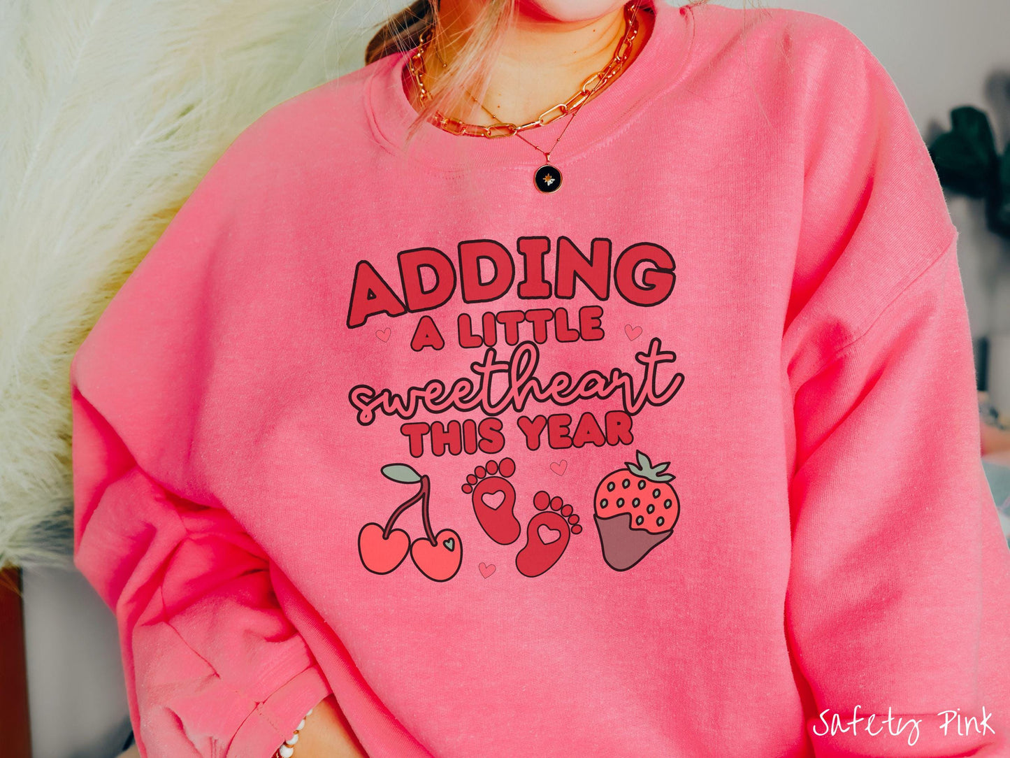 Share your exciting news with this vintage Gildan sweatshirt featuring the phrase “Adding a little sweetheart this year” in bold red font, paired with two red baby footprints framed by two red cherries and a chocolate-covered strawberry.