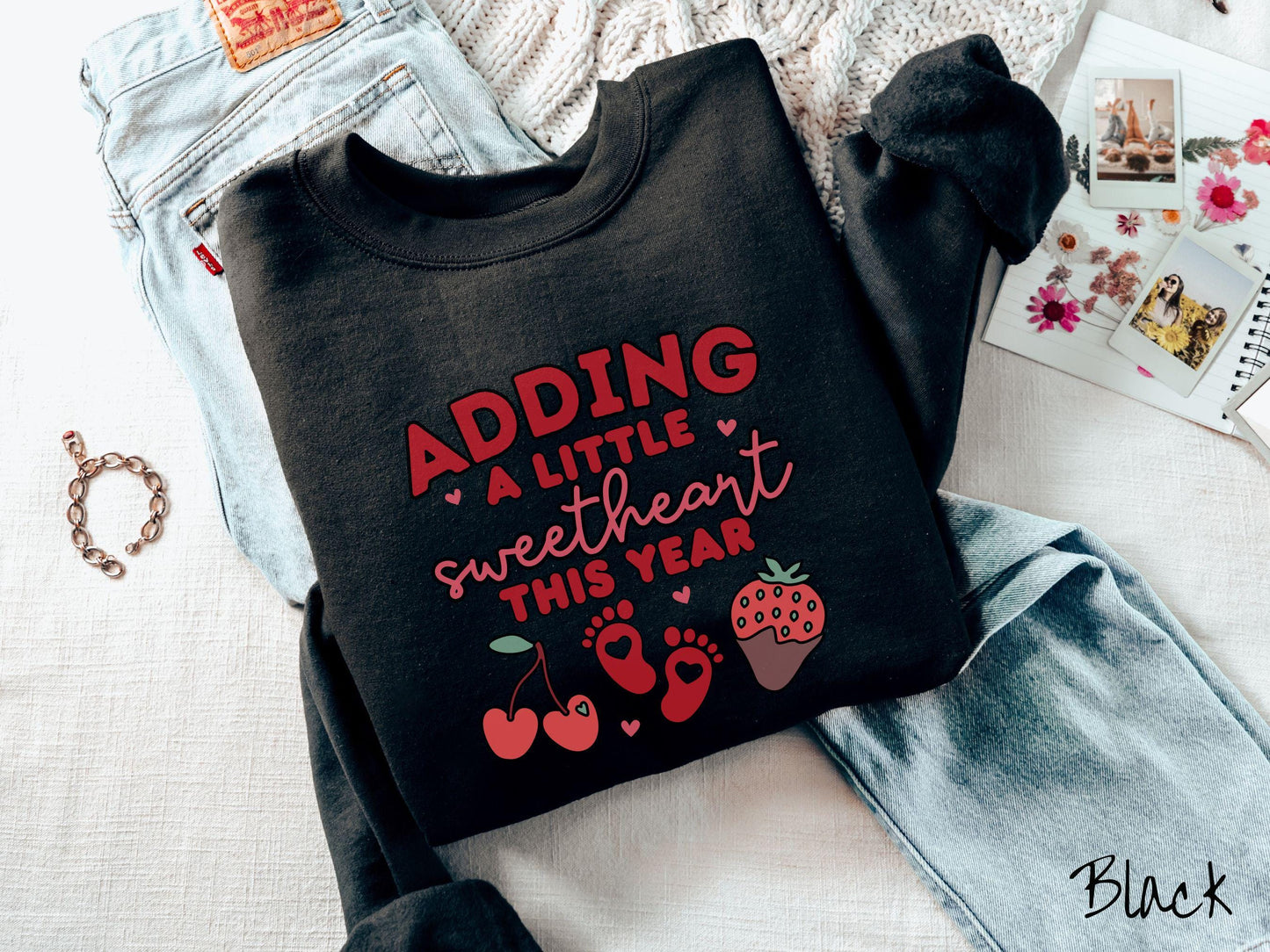 Share your exciting news with this vintage Gildan sweatshirt featuring the phrase “Adding a little sweetheart this year” in bold red font, paired with two red baby footprints framed by two red cherries and a chocolate-covered strawberry.