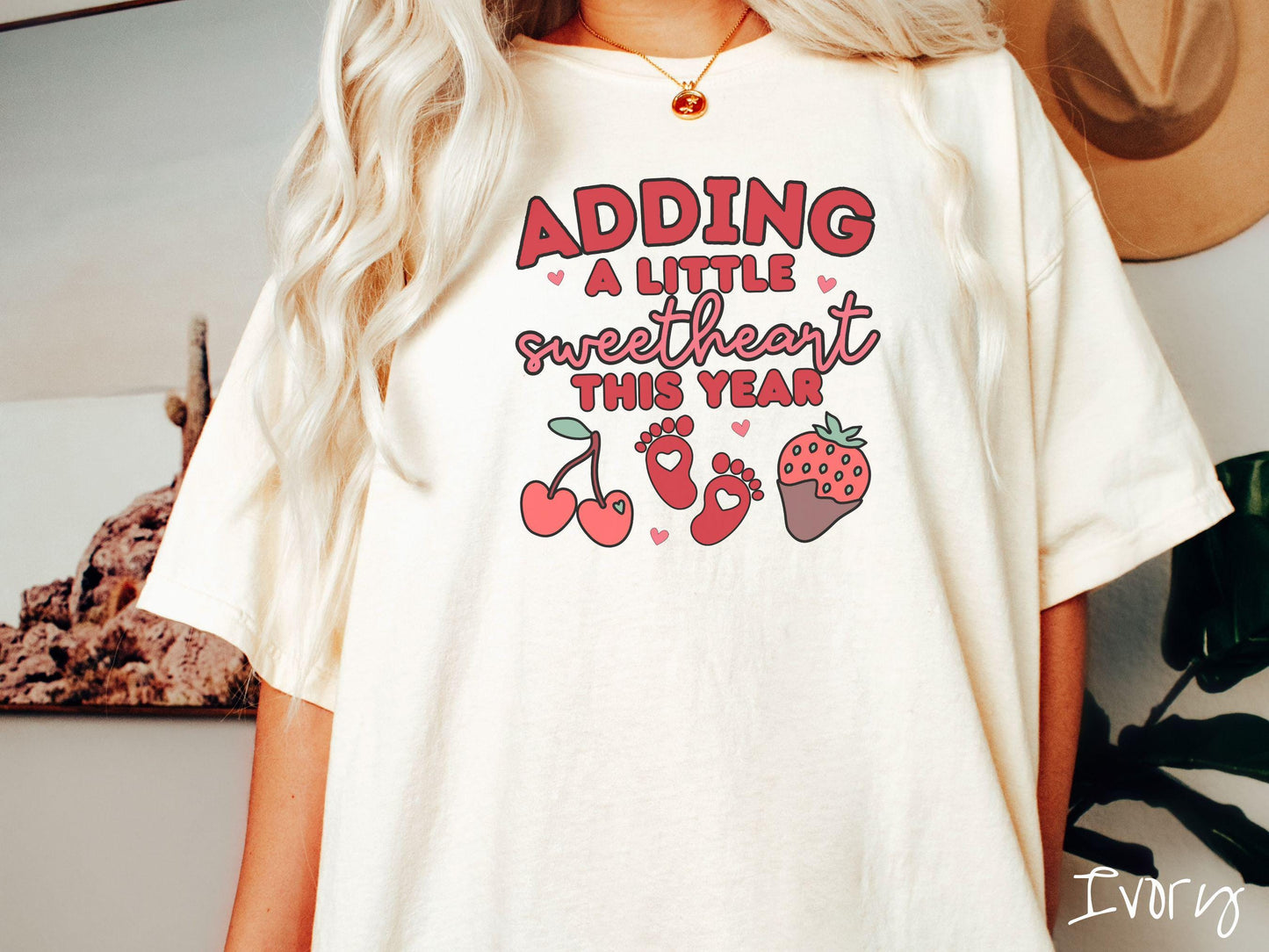 Share your exciting news with this vintage Comfort Colors t-shirt featuring the phrase “Adding a little sweetheart this year” in bold red font, paired with two red baby footprints framed by two red cherries and a chocolate-covered strawberry.