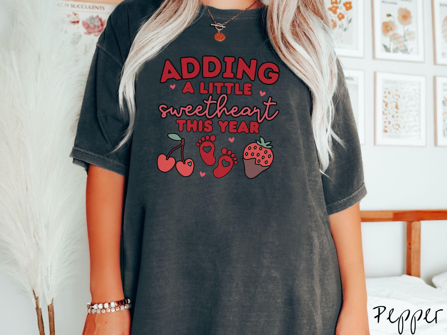 Share your exciting news with this vintage Comfort Colors t-shirt featuring the phrase “Adding a little sweetheart this year” in bold red font, paired with two red baby footprints framed by two red cherries and a chocolate-covered strawberry.