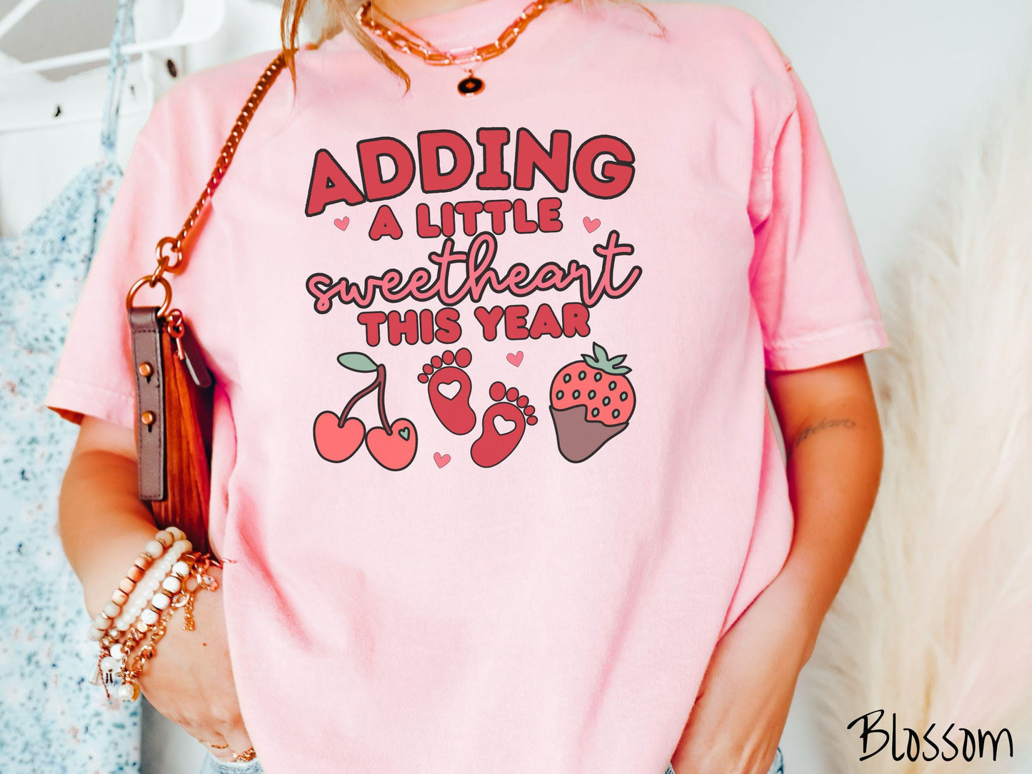 Share your exciting news with this vintage Comfort Colors t-shirt featuring the phrase “Adding a little sweetheart this year” in bold red font, paired with two red baby footprints framed by two red cherries and a chocolate-covered strawberry.