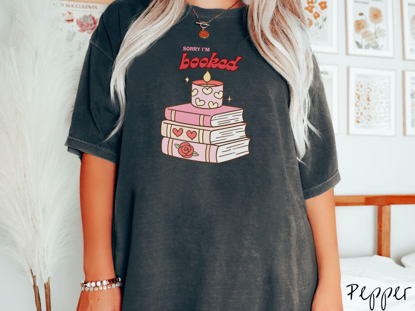 A vintage Comfort Colors t-shirt featuring the playful phrase “Sorry I’m Booked” in pink and red font, completed with a stack of three pink and purple books adorned with red roses and hearts on their spines, topped with a lit candle.