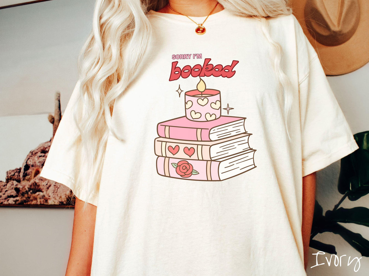 A vintage Comfort Colors t-shirt featuring the playful phrase “Sorry I’m Booked” in pink and red font, completed with a stack of three pink and purple books adorned with red roses and hearts on their spines, topped with a lit candle.