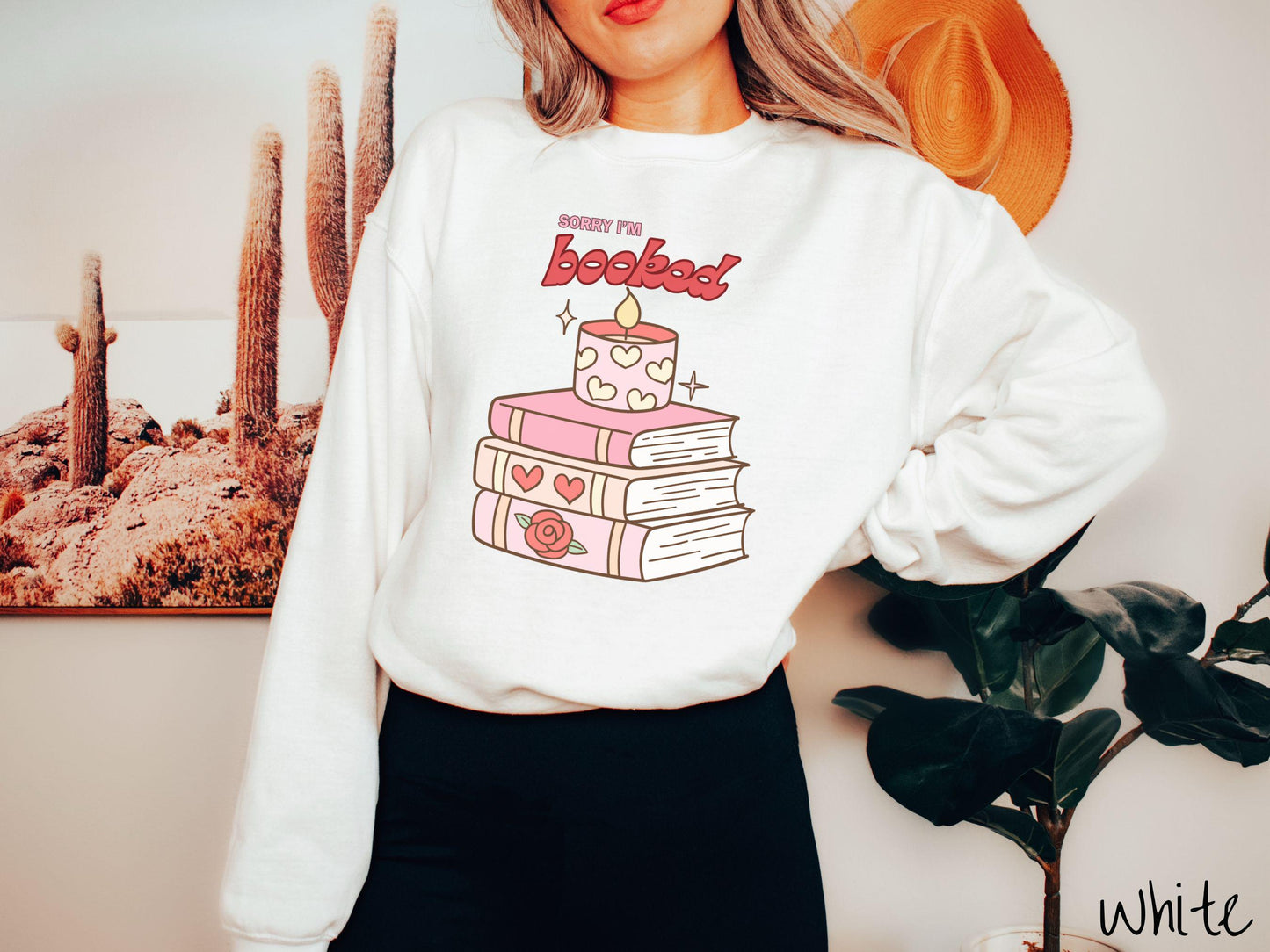A vintage Gildan sweatshirt featuring the playful phrase “Sorry I’m Booked” in pink and red font, completed with a stack of three pink and purple books adorned with red roses and hearts on their spines, topped with a lit candle.