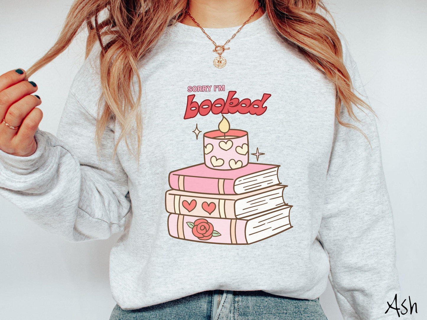 A vintage Gildan sweatshirt featuring the playful phrase “Sorry I’m Booked” in pink and red font, completed with a stack of three pink and purple books adorned with red roses and hearts on their spines, topped with a lit candle.