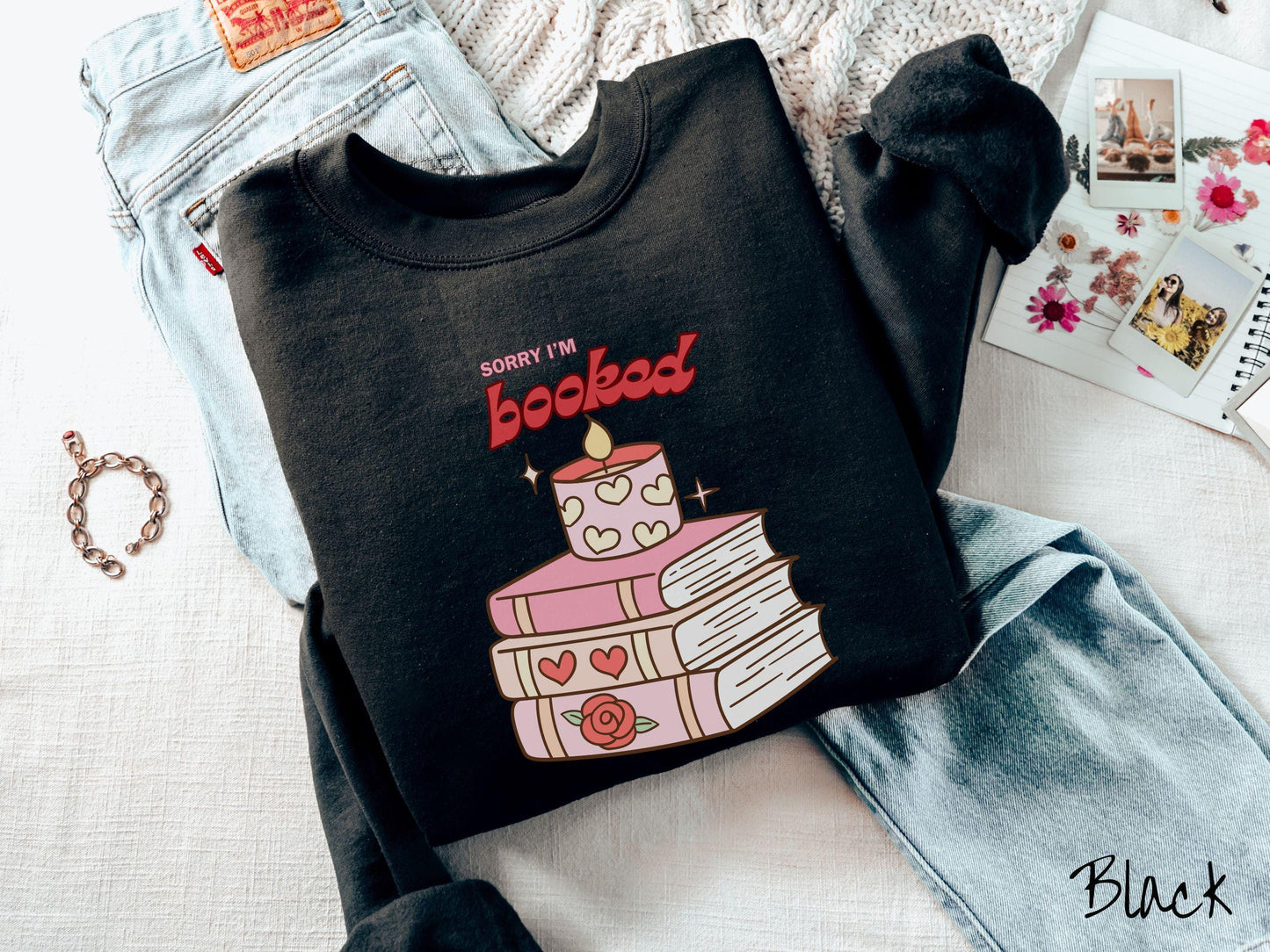 A vintage Gildan sweatshirt featuring the playful phrase “Sorry I’m Booked” in pink and red font, completed with a stack of three pink and purple books adorned with red roses and hearts on their spines, topped with a lit candle.