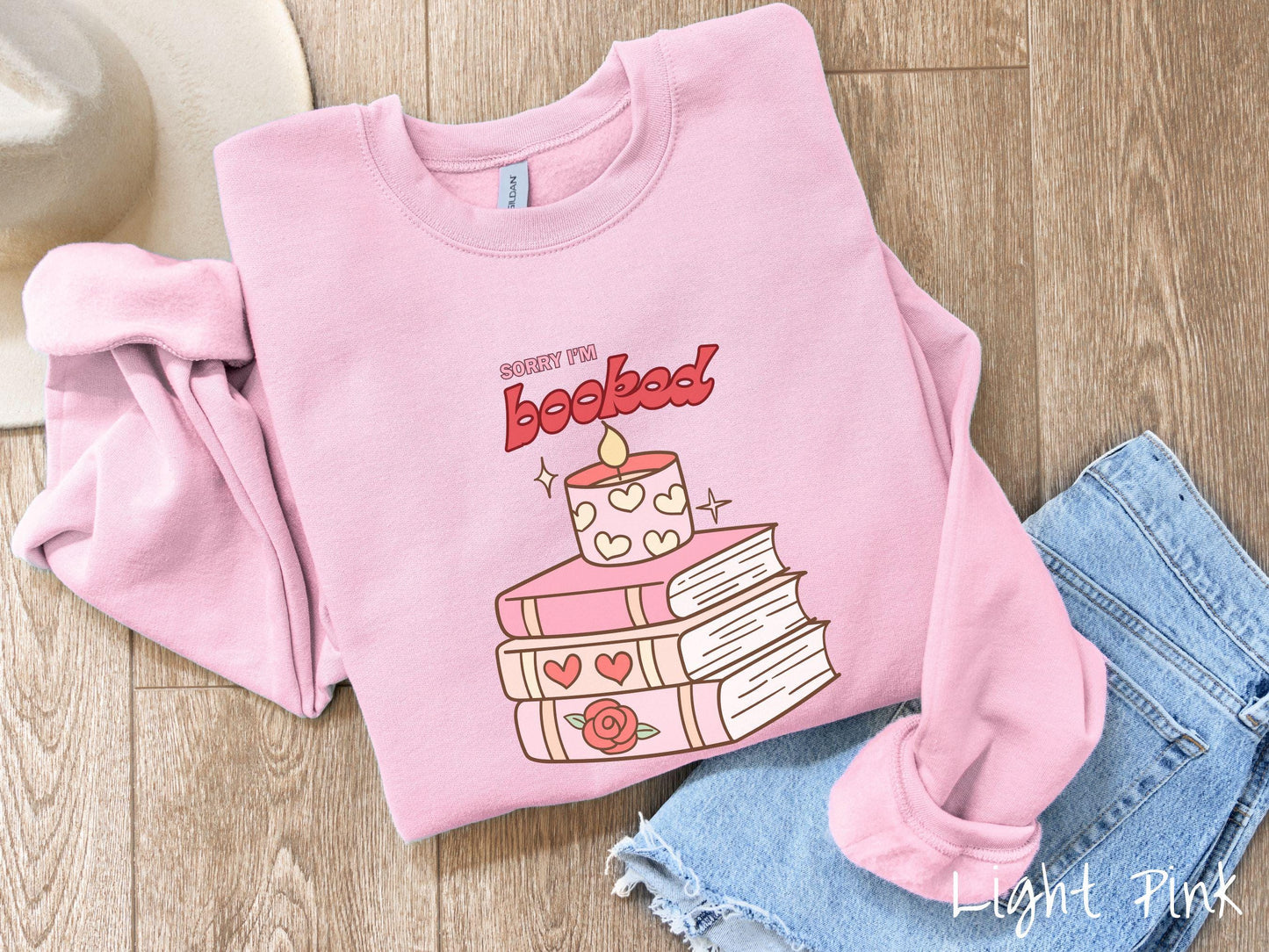 A vintage Gildan sweatshirt featuring the playful phrase “Sorry I’m Booked” in pink and red font, completed with a stack of three pink and purple books adorned with red roses and hearts on their spines, topped with a lit candle.