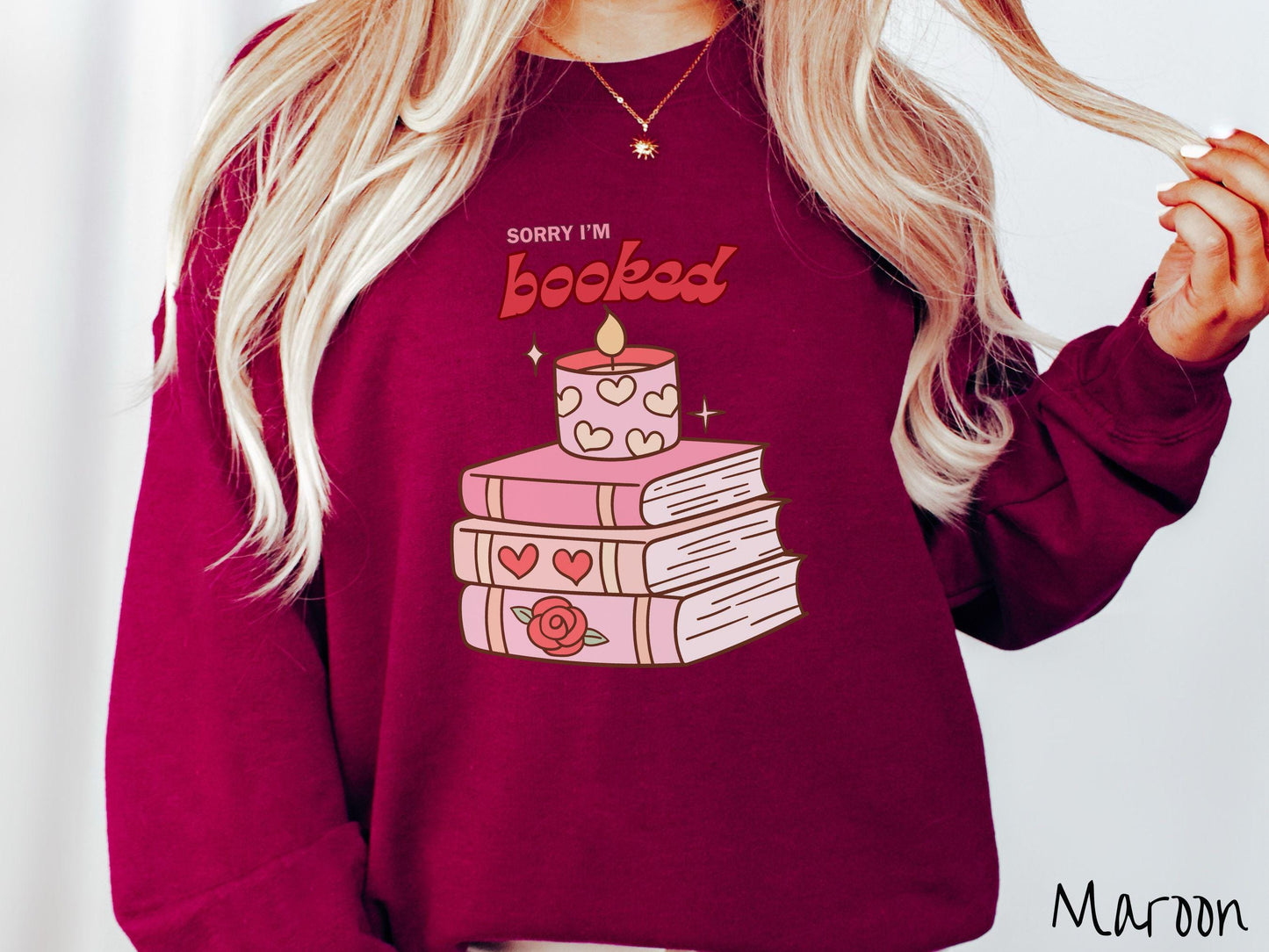 A vintage Gildan sweatshirt featuring the playful phrase “Sorry I’m Booked” in pink and red font, completed with a stack of three pink and purple books adorned with red roses and hearts on their spines, topped with a lit candle.