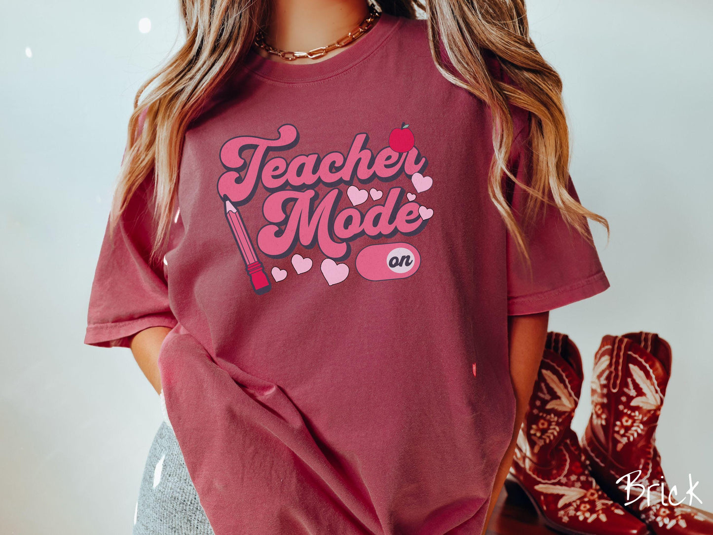 A vintage Comfort Colors t-shirt featuring the phrase “Teacher Mode” in playful pink font, an “On” button toggled to the “On” position, and with charming details like a red pencil, a red apple, and pink hearts sprinkled throughout.