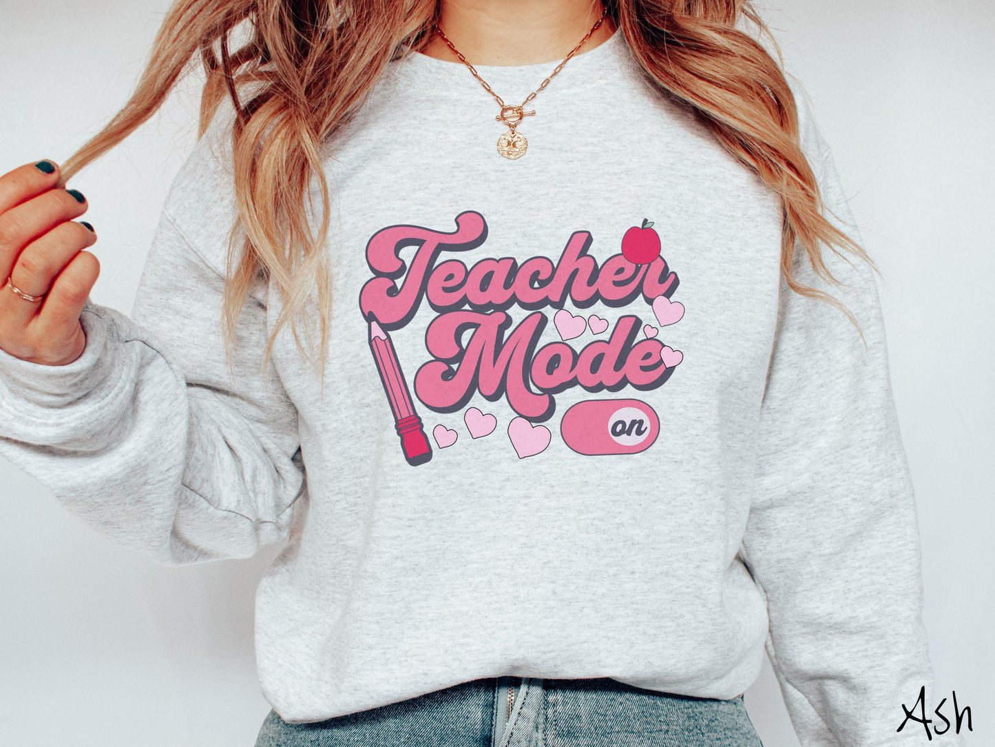 A vintage Gildan sweathirt featuring the phrase “Teacher Mode” in playful pink font, an “On” button toggled to the “On” position, and with charming details like a red pencil, a red apple, and pink hearts sprinkled throughout.