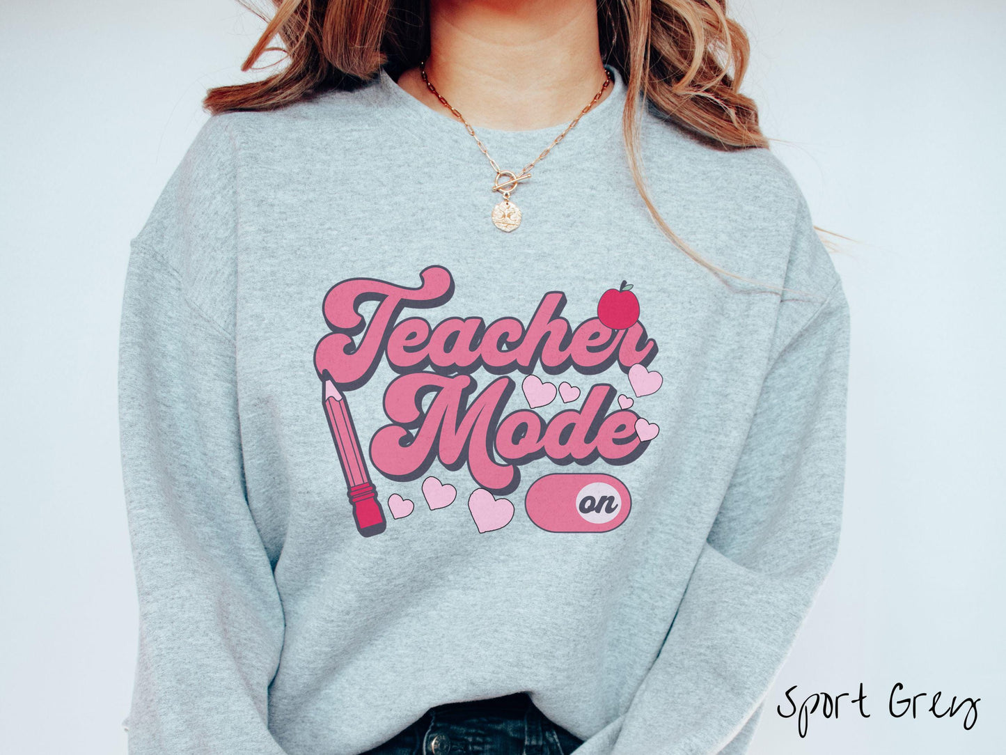 A vintage Gildan sweathirt featuring the phrase “Teacher Mode” in playful pink font, an “On” button toggled to the “On” position, and with charming details like a red pencil, a red apple, and pink hearts sprinkled throughout.