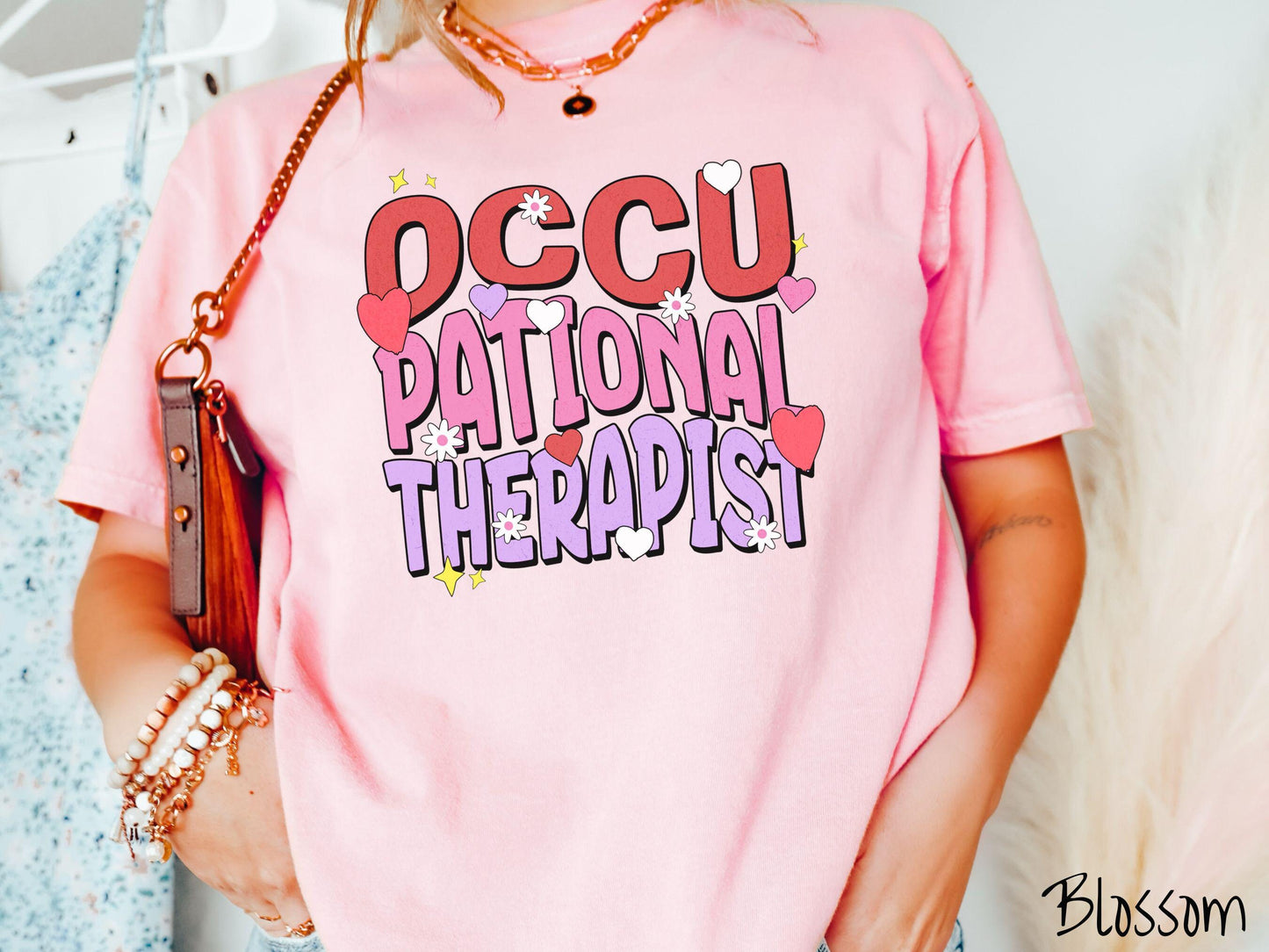 A stylish Comfort Colors t-shirt featuring the words "Occupational Therapist" arranged vertically in bold red, pink, and purple font, this design is enhanced with colorful hearts, flowers, and stars for a playful, uplifting touch.
