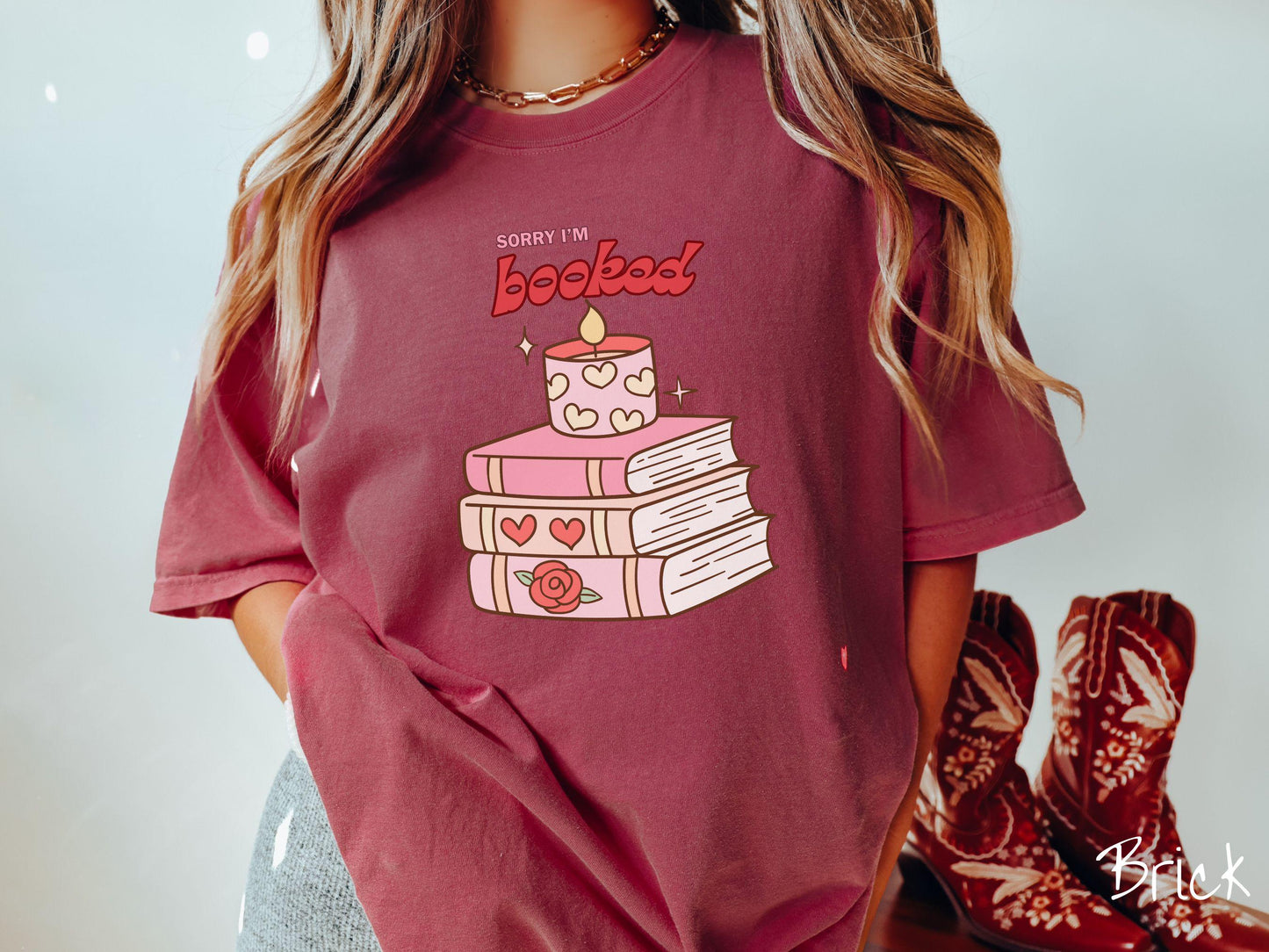 A vintage Comfort Colors t-shirt featuring the playful phrase “Sorry I’m Booked” in pink and red font, completed with a stack of three pink and purple books adorned with red roses and hearts on their spines, topped with a lit candle.