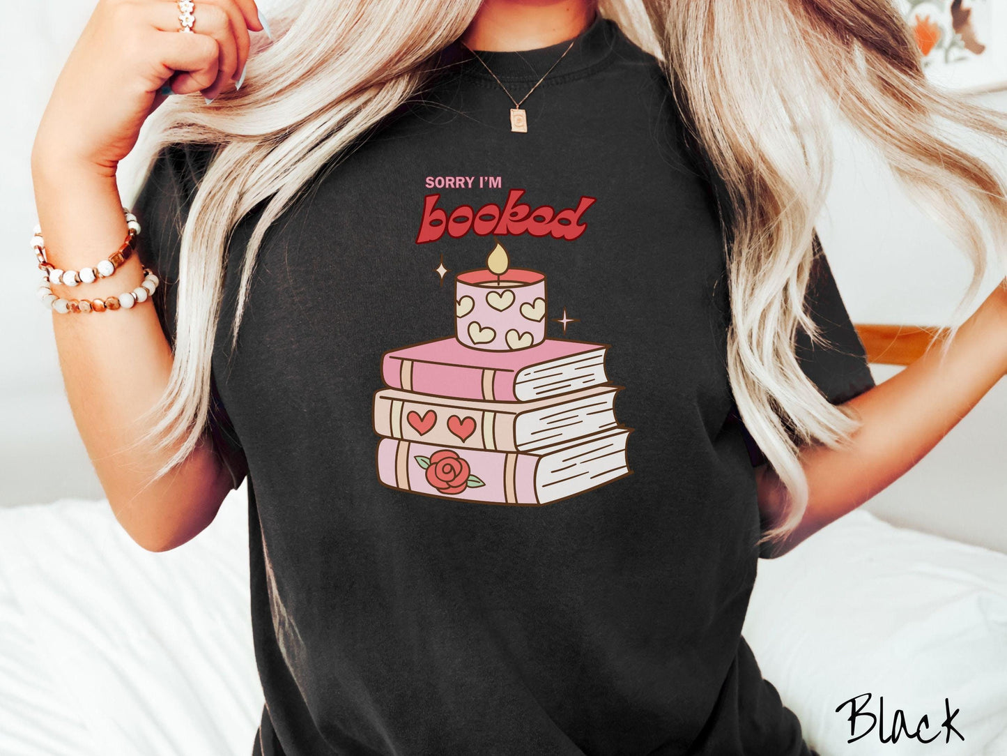 A vintage Comfort Colors t-shirt featuring the playful phrase “Sorry I’m Booked” in pink and red font, completed with a stack of three pink and purple books adorned with red roses and hearts on their spines, topped with a lit candle.
