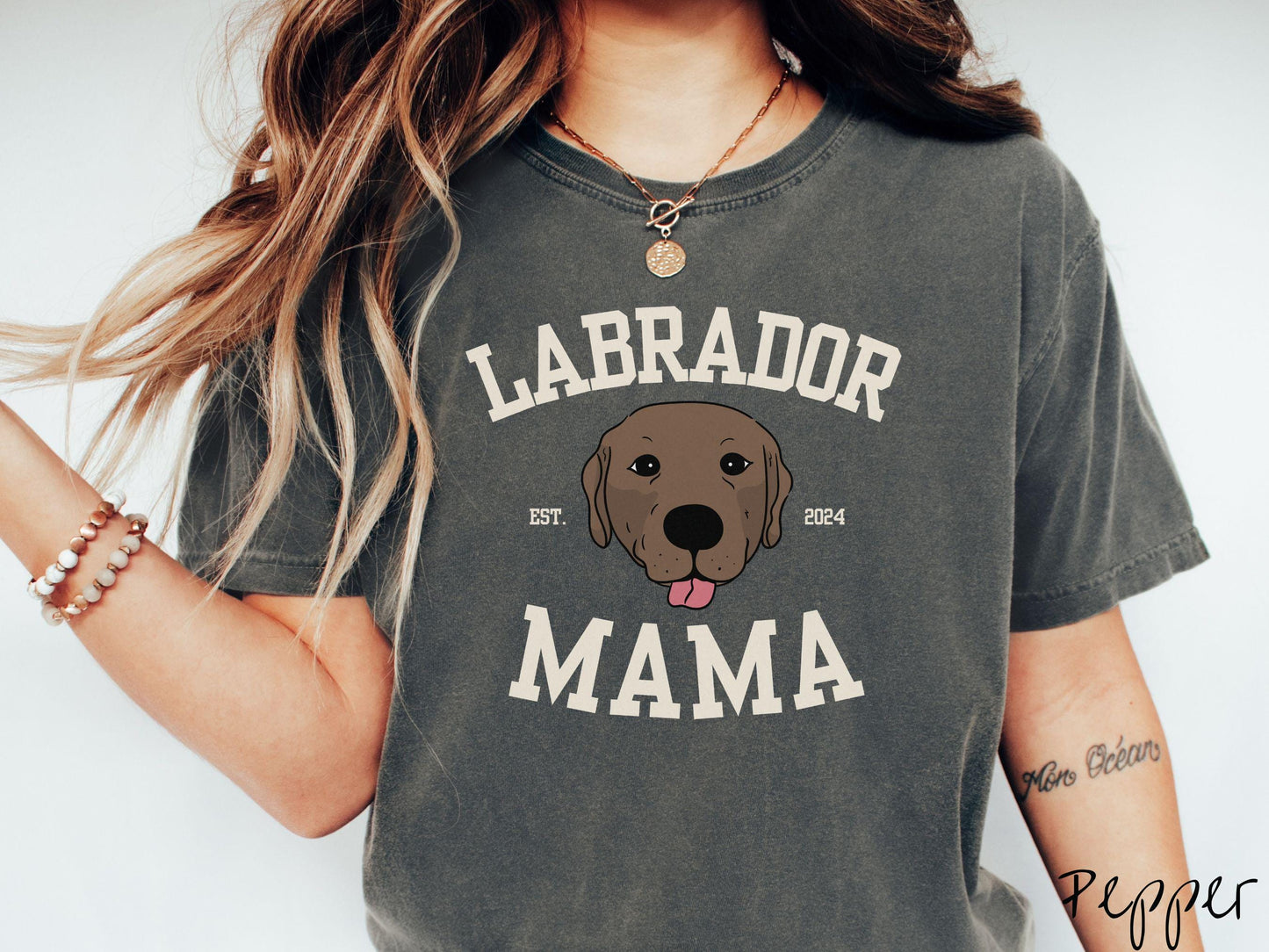 A vintage Comfort Colors t-shirt featuring bold block lettering that reads 'Labrador Mama.' At the center of the design, a playful chocolate Labrador with its tongue out steals the spotlight, framed by the stylish text 'EST. 2024.'