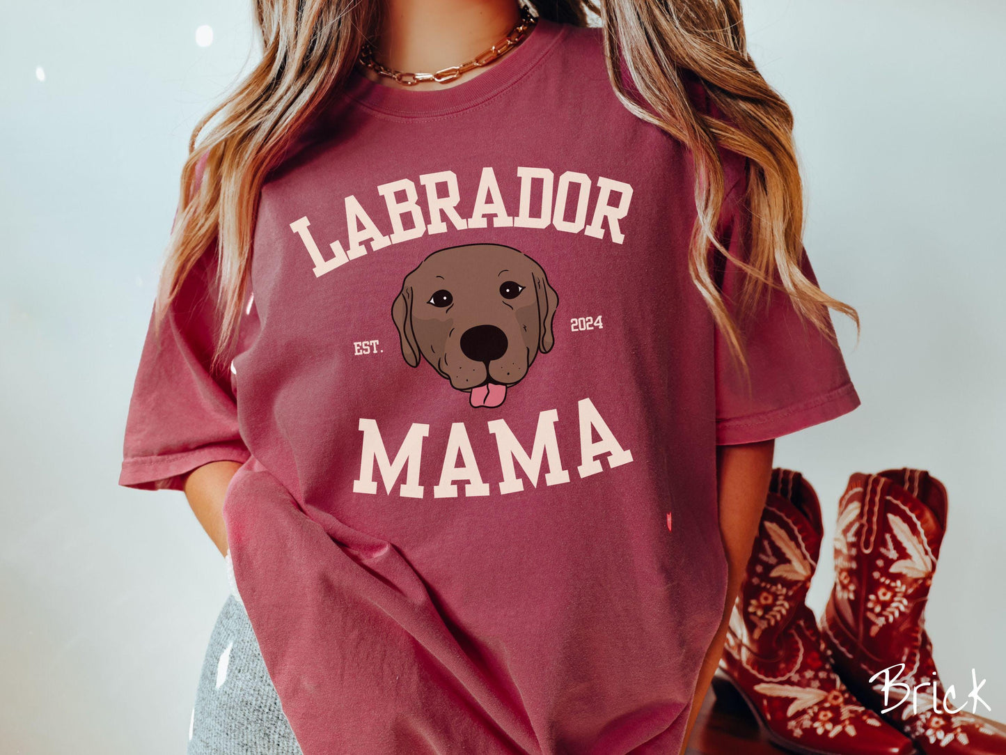 A vintage Comfort Colors t-shirt featuring bold block lettering that reads 'Labrador Mama.' At the center of the design, a playful chocolate Labrador with its tongue out steals the spotlight, framed by the stylish text 'EST. 2024.'