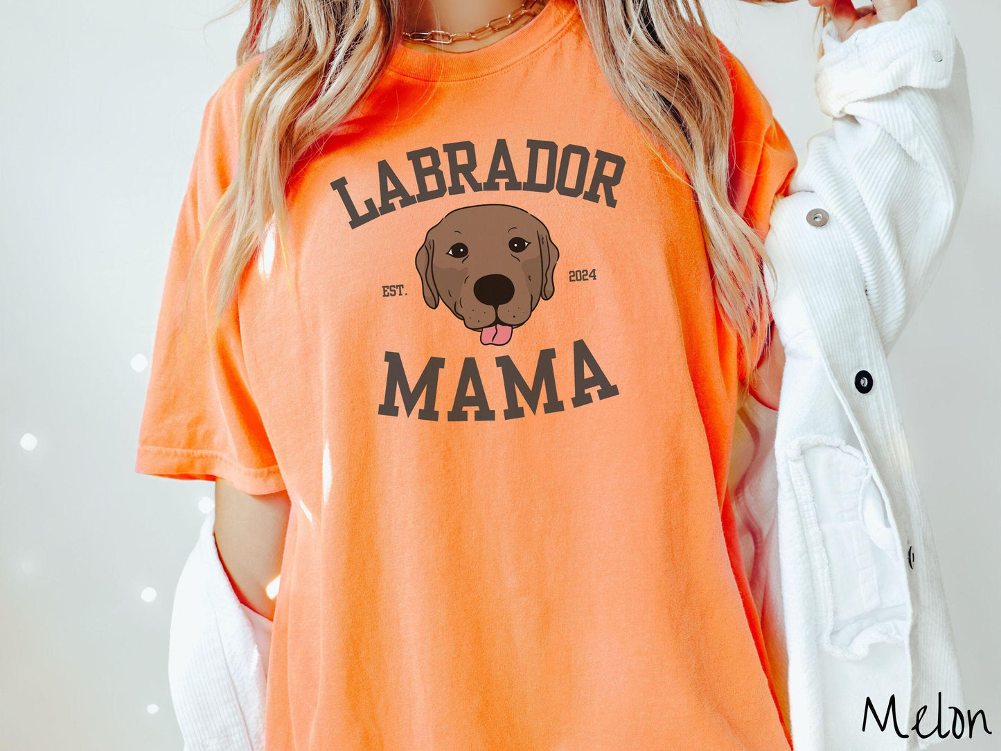 A vintage Comfort Colors t-shirt featuring bold block lettering that reads 'Labrador Mama.' At the center of the design, a playful chocolate Labrador with its tongue out steals the spotlight, framed by the stylish text 'EST. 2024.'
