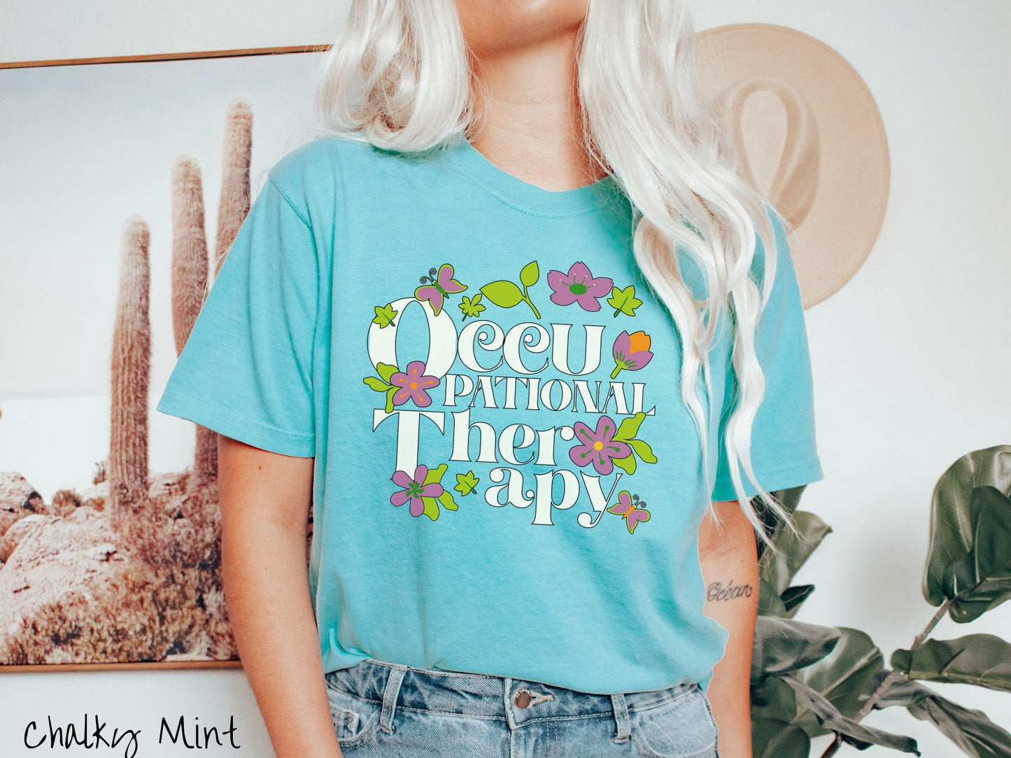 A vintage, chalky mint colored Comfort Colors t-shirt with the text Occupational Therapy and featuring green leaves, purple and orange flowers, and colorful butterflies in the background.
