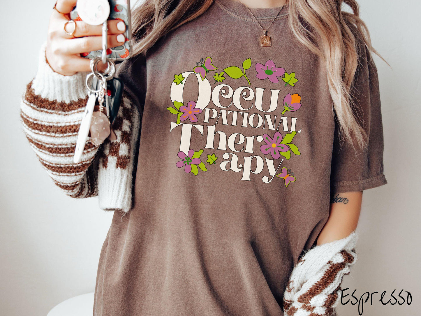 A vintage, espresso colored Comfort Colors t-shirt with the text Occupational Therapy and featuring green leaves, purple and orange flowers, and colorful butterflies in the background.