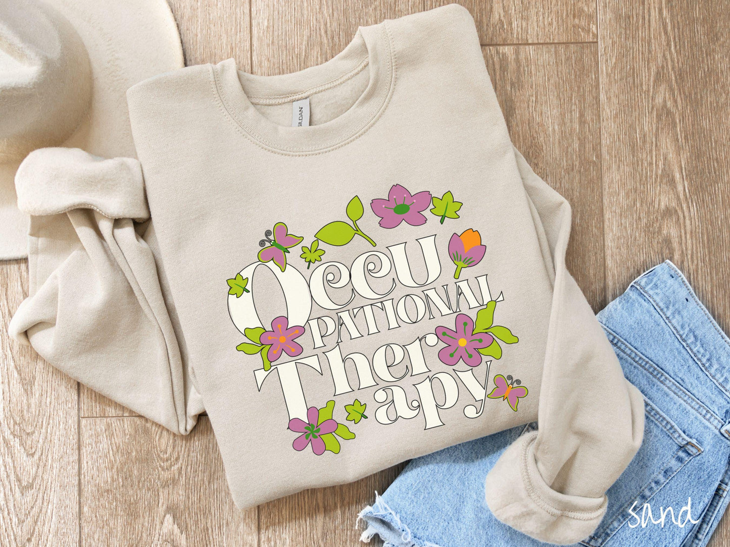 A vintage, sand colored Gildan sweatshirt with the text Occupational Therapy and featuring green leaves, purple and orange flowers, and colorful butterflies in the background.