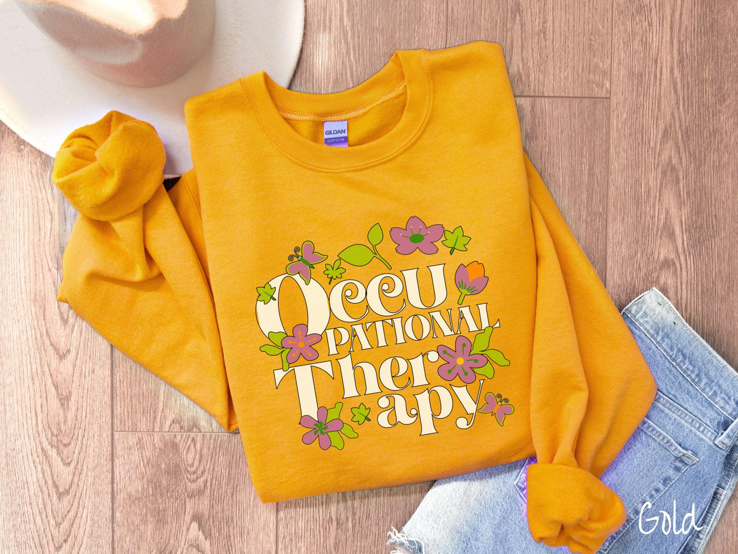 A vintage, gold colored Gildan sweatshirt with the text Occupational Therapy and featuring green leaves, purple and orange flowers, and colorful butterflies in the background.