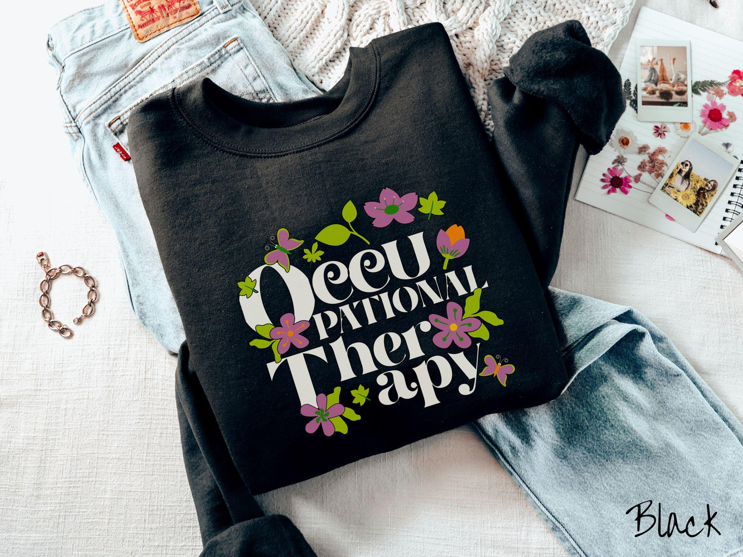 A vintage, black colored Gildan sweatshirt with the text Occupational Therapy and featuring green leaves, purple and orange flowers, and colorful butterflies in the background.