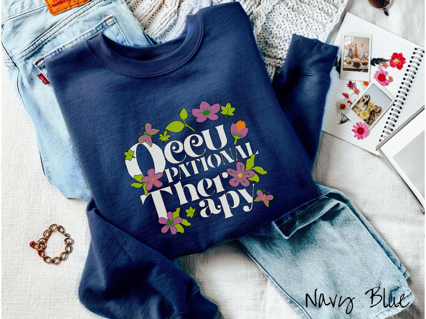 A vintage, navy blue colored Gildan sweatshirt with the text Occupational Therapy and featuring green leaves, purple and orange flowers, and colorful butterflies in the background.