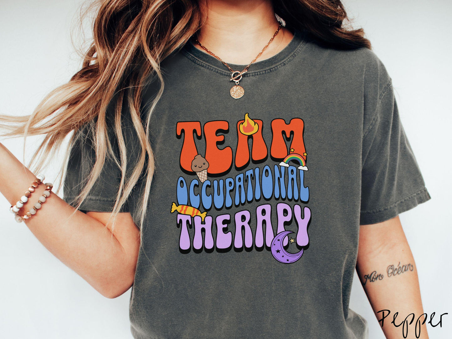 A vintage, pepper colored Comfort Colors t-shirt with the text "Team Occupational Therapy" surrounded by playful elements like a chocolate ice cream cone, a blazing flame, a colorful rainbow, a piece of candy, and a serene moon.